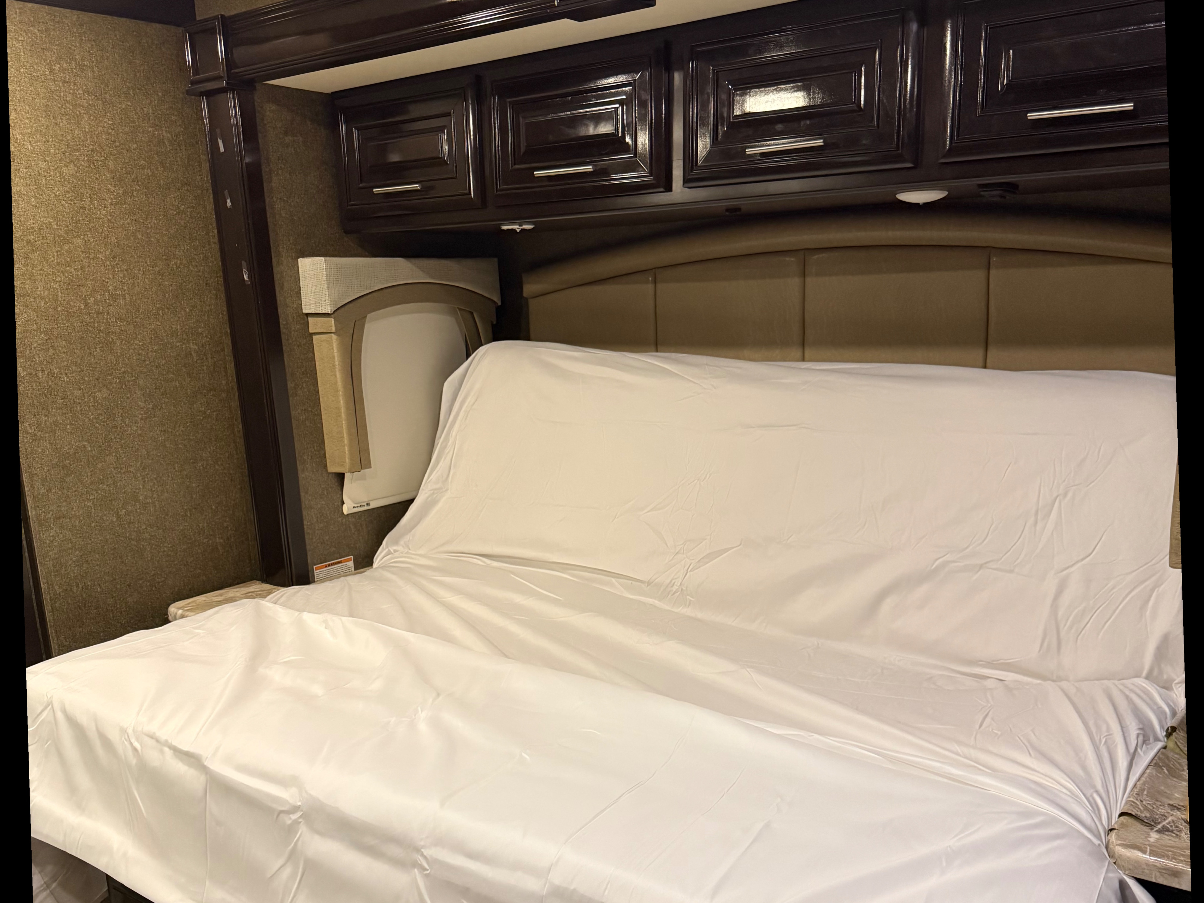 RV rental bedroom features a cozy bed with white sheets, padded headboard, dark cabinets, and textured walls.. Thor Miramar 2019