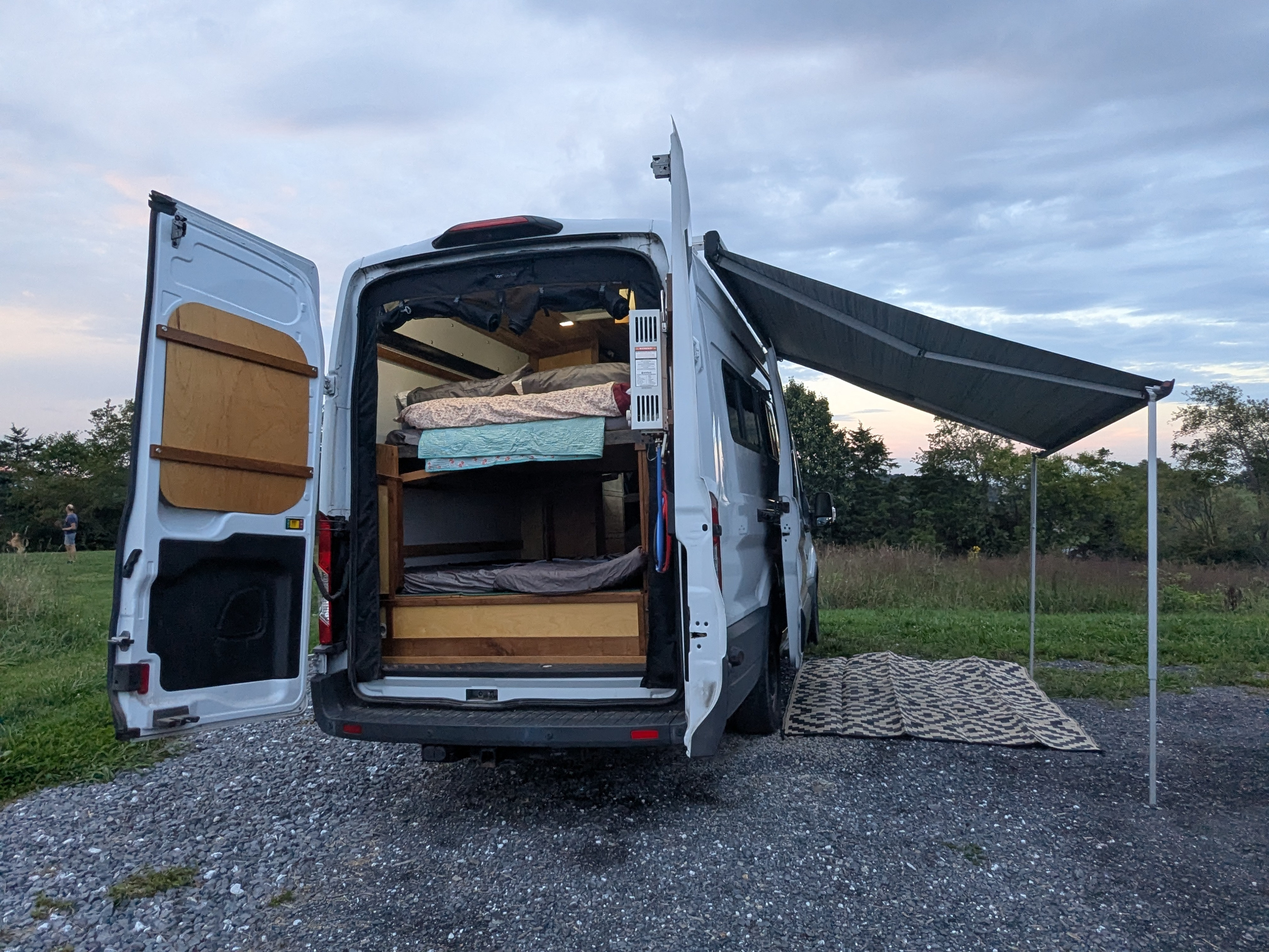 This RV rental's open back reveals bunk beds, with an extended awning and mat set up for an outdoor adventure.. Ford Transit custom 2018