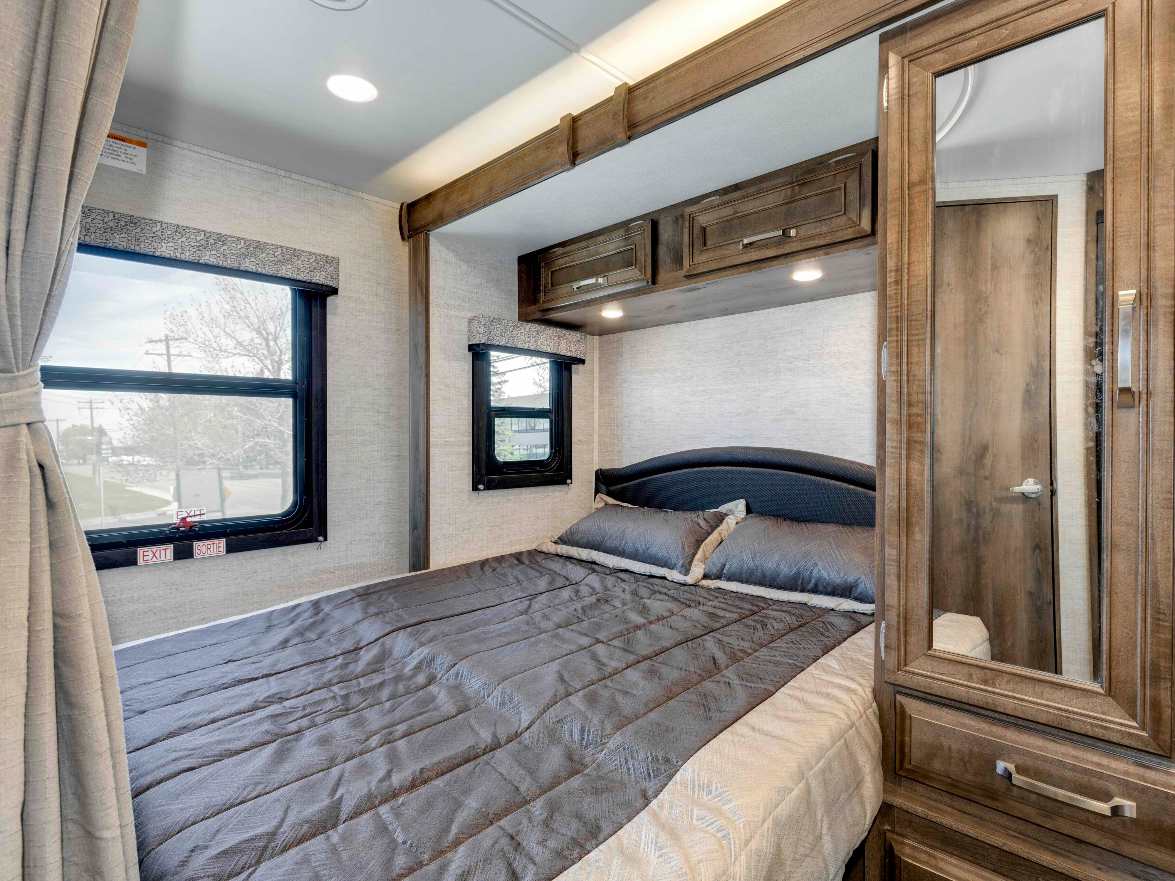 RV rental bedroom with a dark grey bed, overhead cabinets, two windows with blinds, and a mirrored wardrobe.. Jayco Melbourne 2022