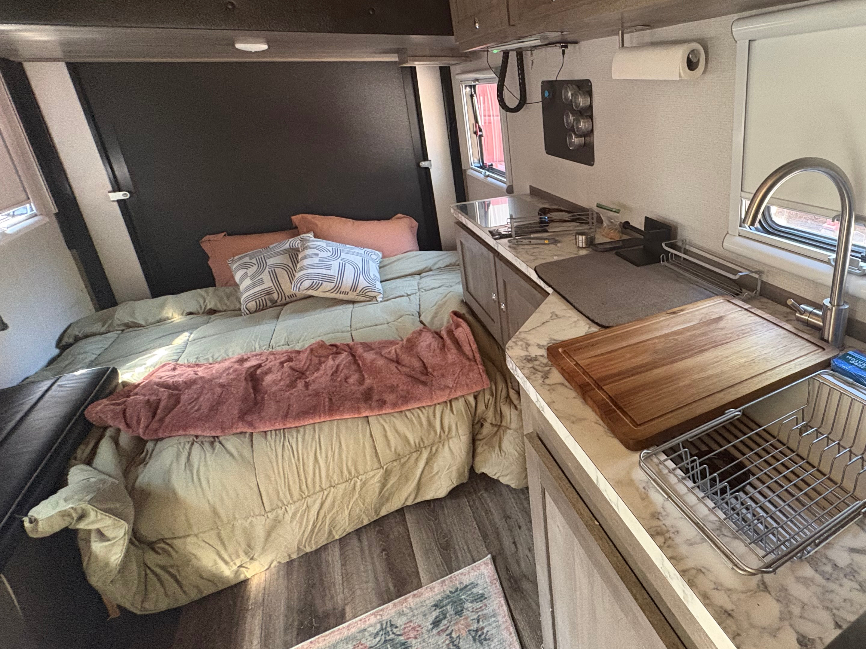 Cozy RV rental interior. A comfortable bed with pillows is on the left, next to a compact kitchen with a sink, cutting board, and dish rack.. Imperial Outdoors XploreRV 2022