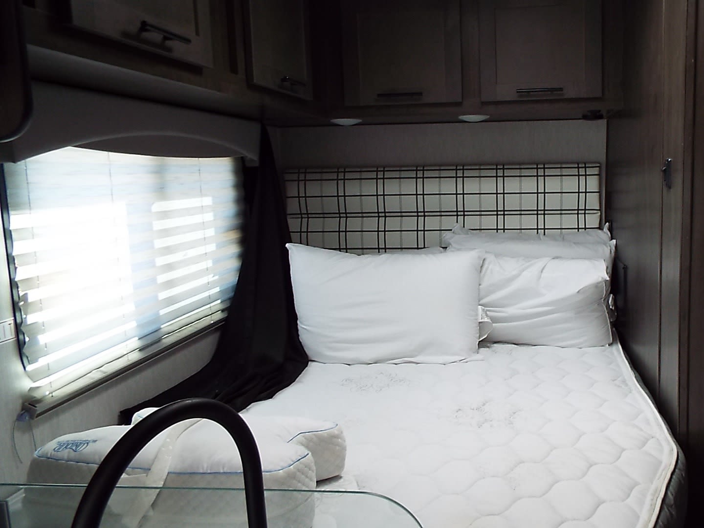 Inside an RV rental, this bedroom features a bed with white pillows, a plaid headboard, overhead cabinets, and a bright window.. Coachmen Ford Transit 2023