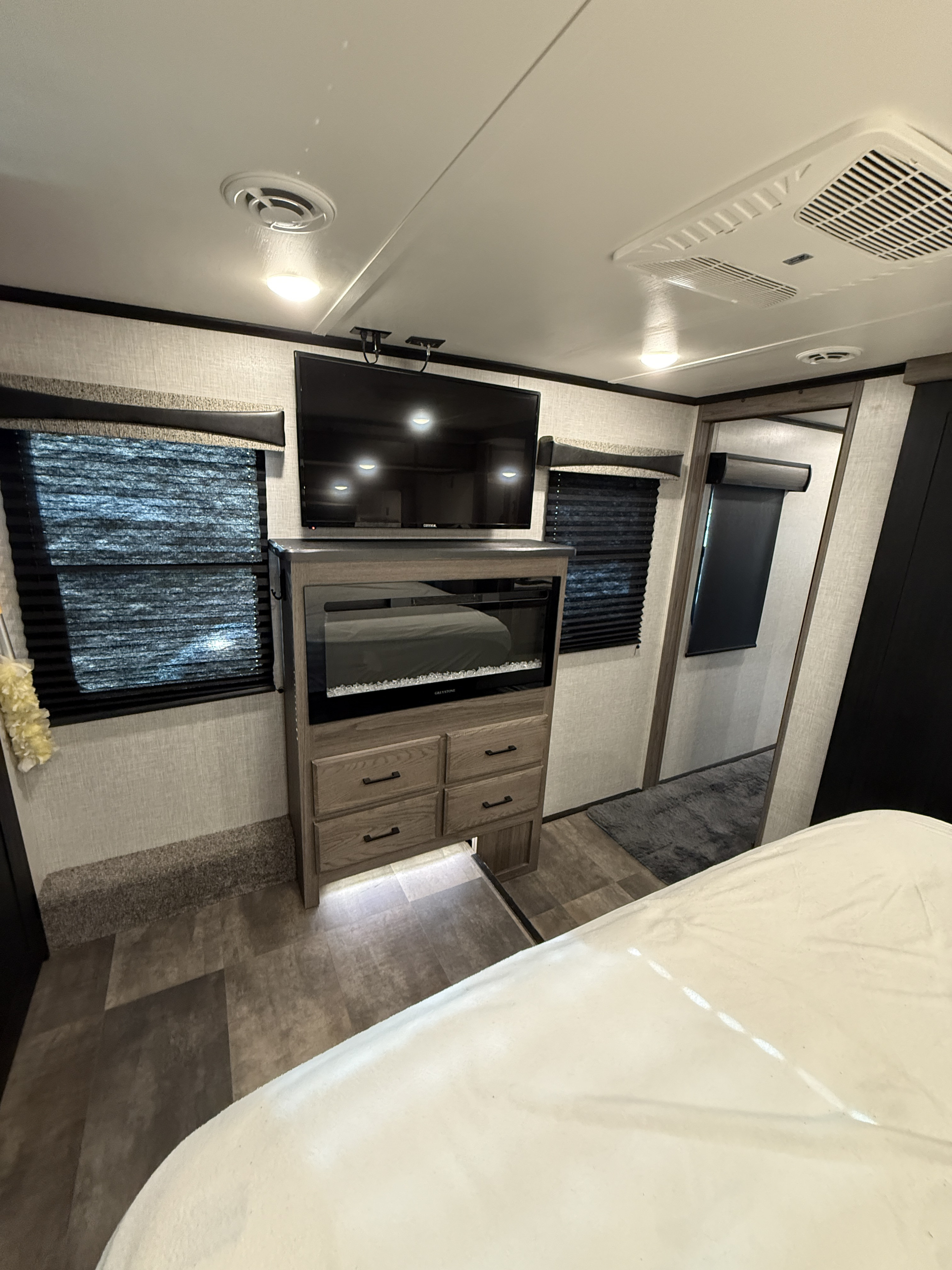 Modern rv rental bedroom with wall-mounted TV, electric fireplace, dresser, window blinds, and bed in foreground.. Crossroads RV Cameo 2022