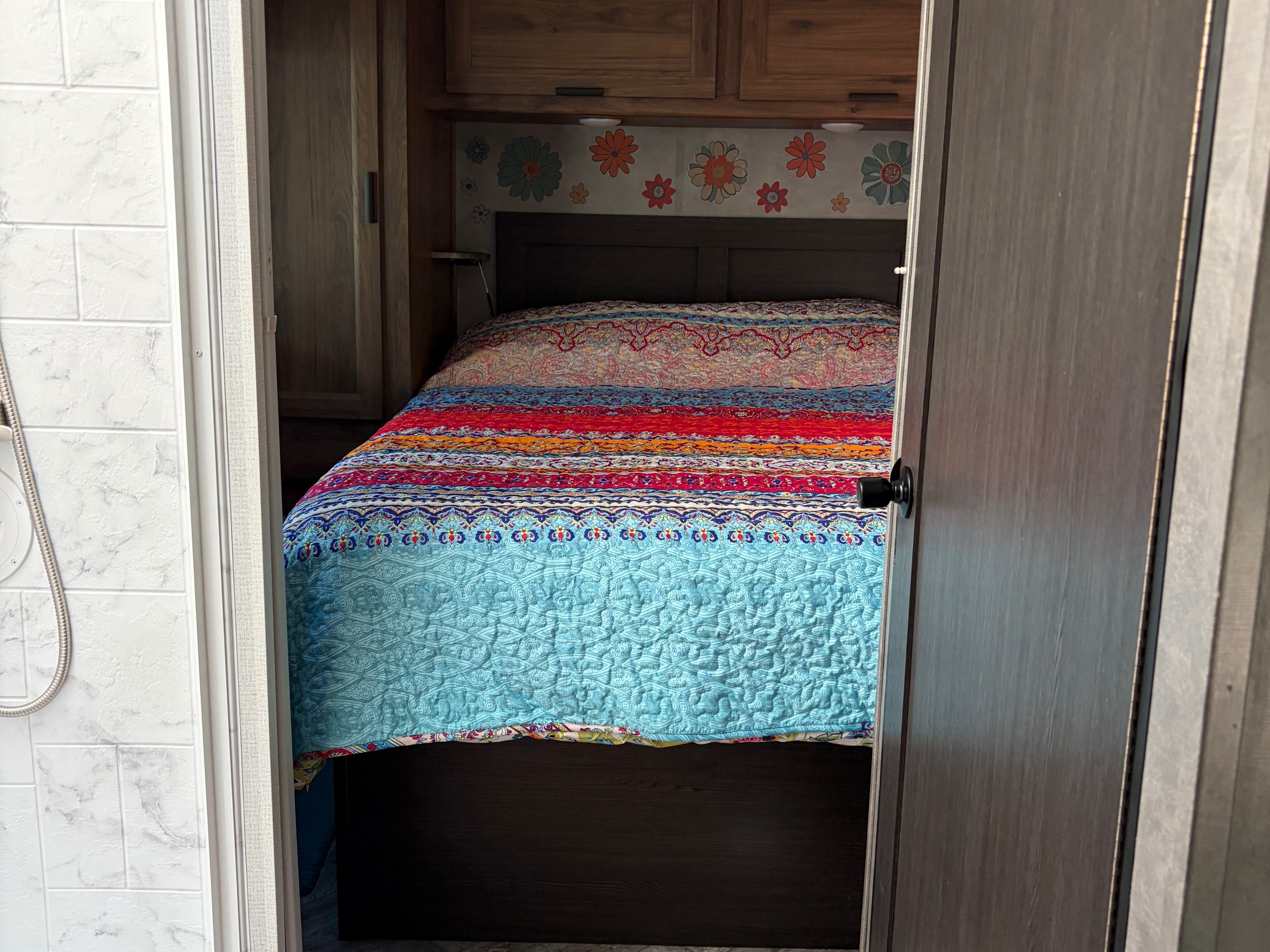 RV rental bedroom: a colorful quilt, wood cabinetry, and floral wall decor are seen through a doorway, with a shower on the left.. Forester LE 2022