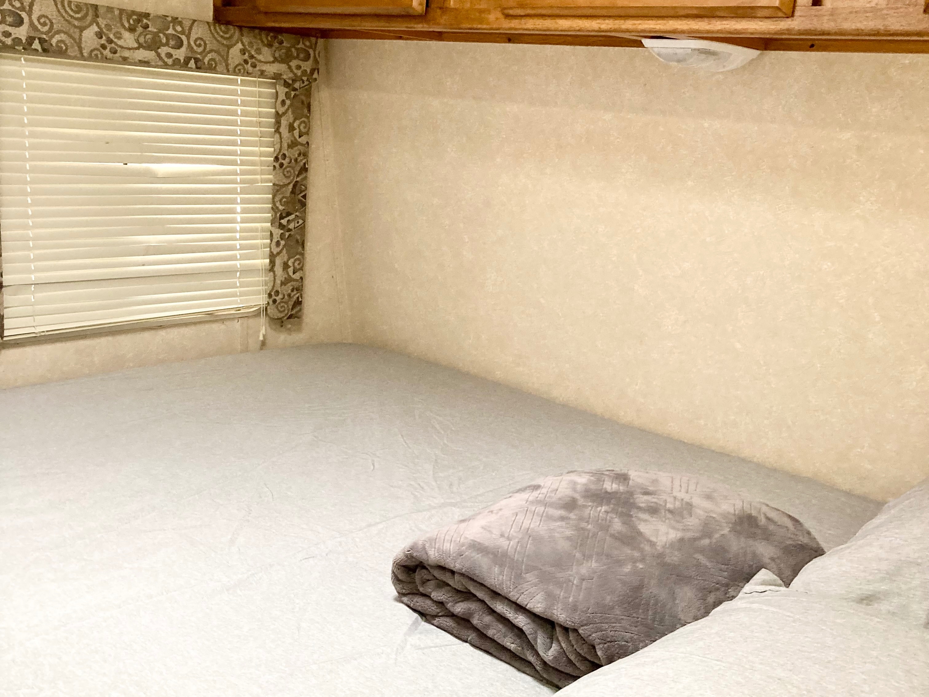 Cozy RV rental bedroom featuring a comfortable bed with light grey bedding, a folded blanket, and window with blinds.. Rage'N Toy Hauler 2006