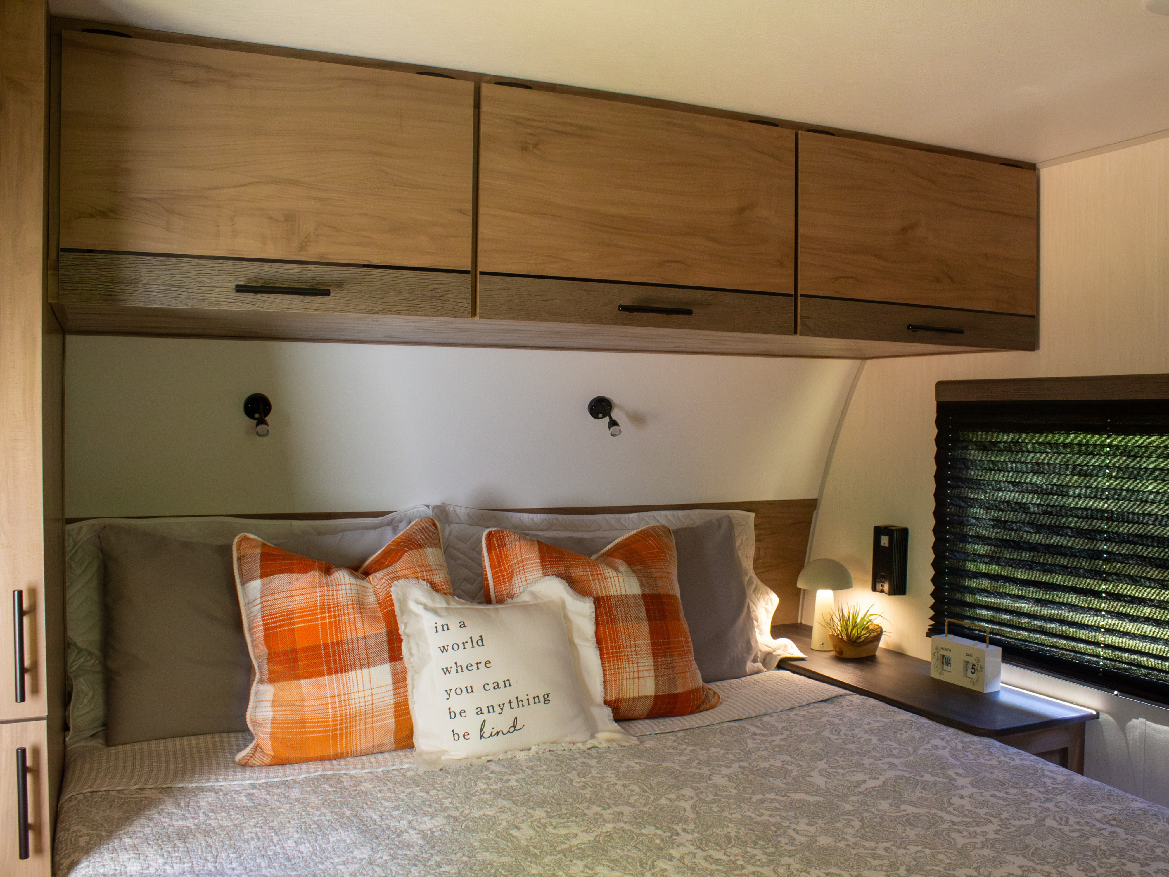 Cozy RV rental bedroom with orange plaid pillows, overhead storage, and a bedside lamp. Perfect for a comfortable road trip.. Forest River Campsite Reserve 2025