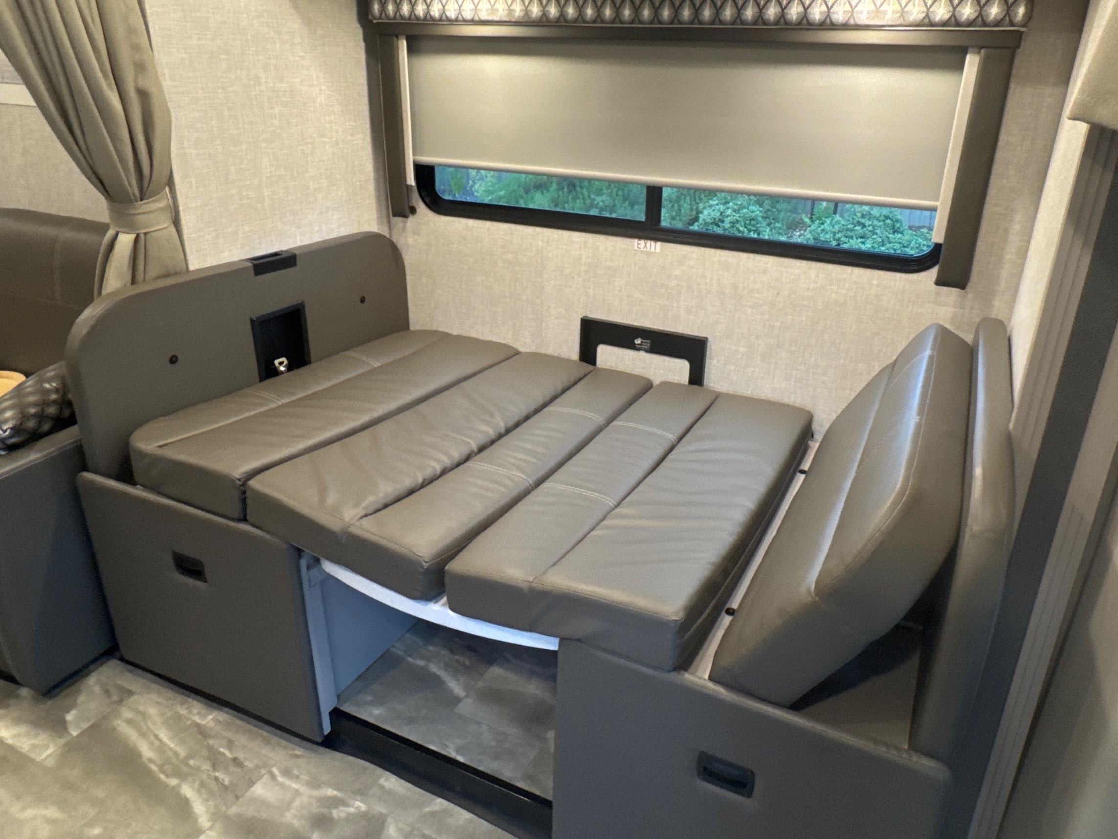 RV rental dinette bed setup. Gray upholstered cushions form the bed, positioned next to a window with a pulled-down blind.. Thor Quantum 2023