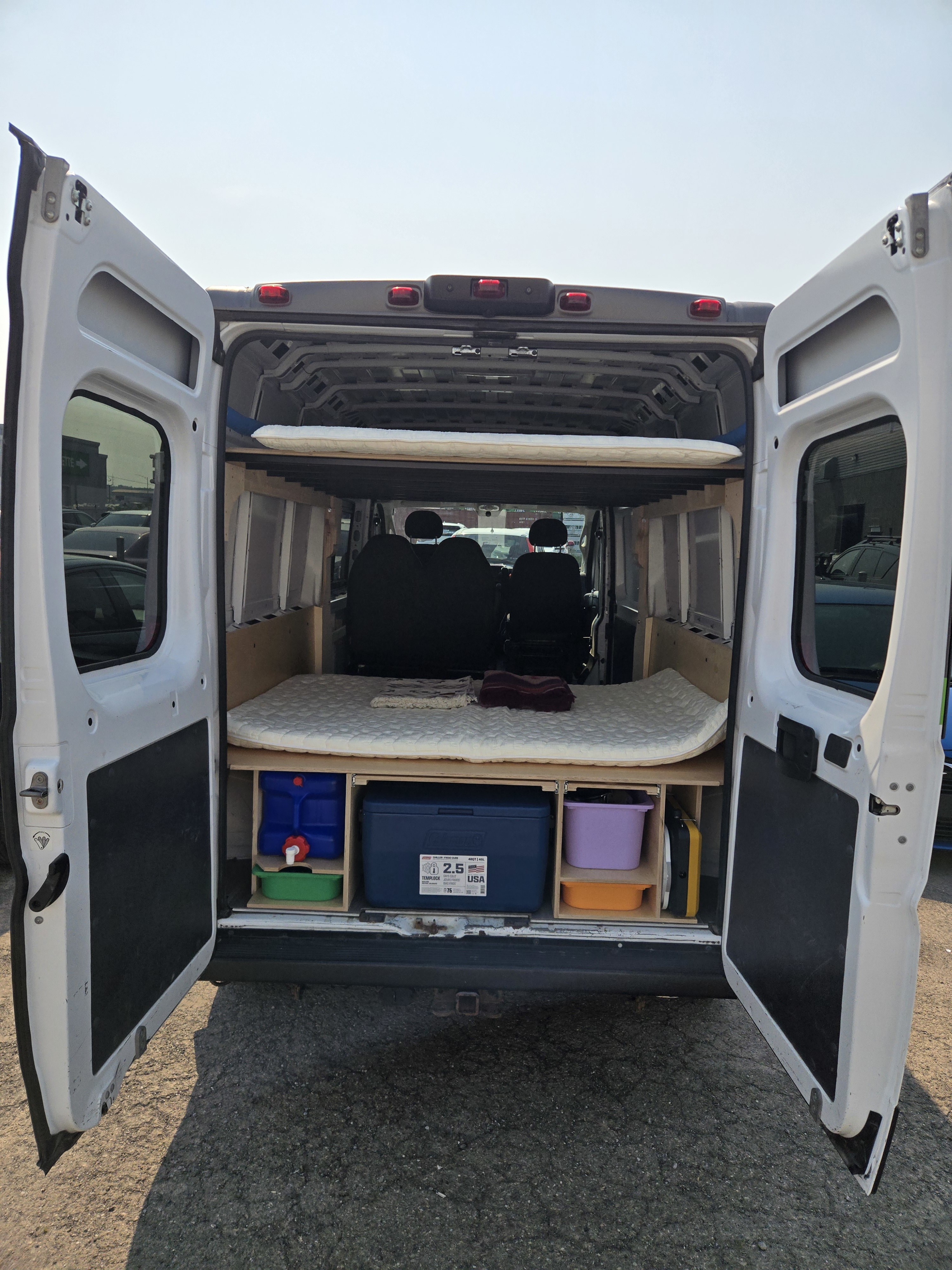 Inside a custom RV rental van: rear doors open to reveal a bed, lower storage with water/cooler, and an upper bunk.. Dodge Promaster 2500 2019