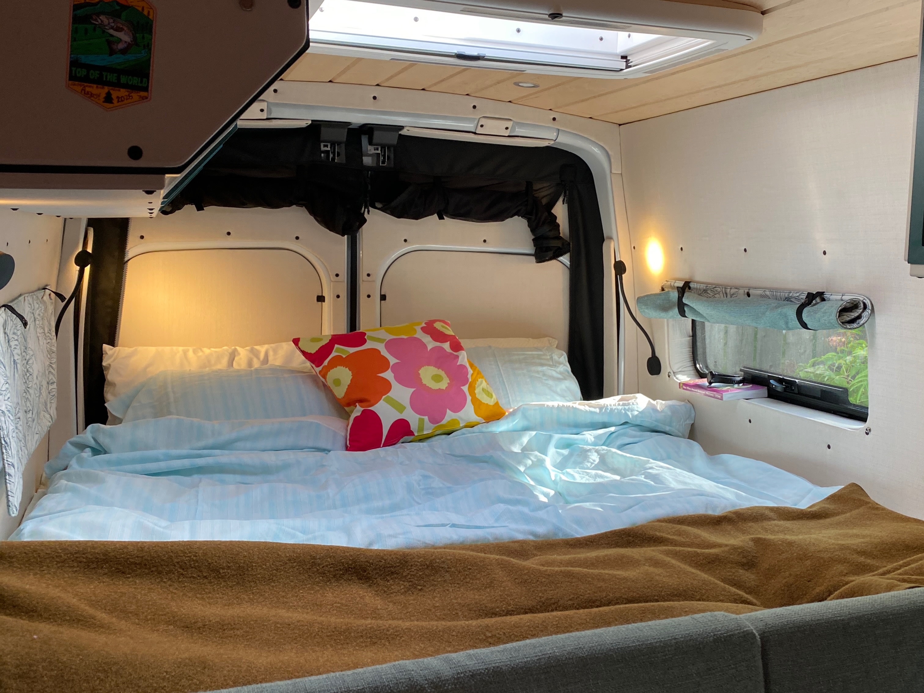 Cozy RV rental bedroom with light blue bedding, colorful pillow, ambient lighting, skylight, and a "Top of the World" sticker.. Ford Transit 2023