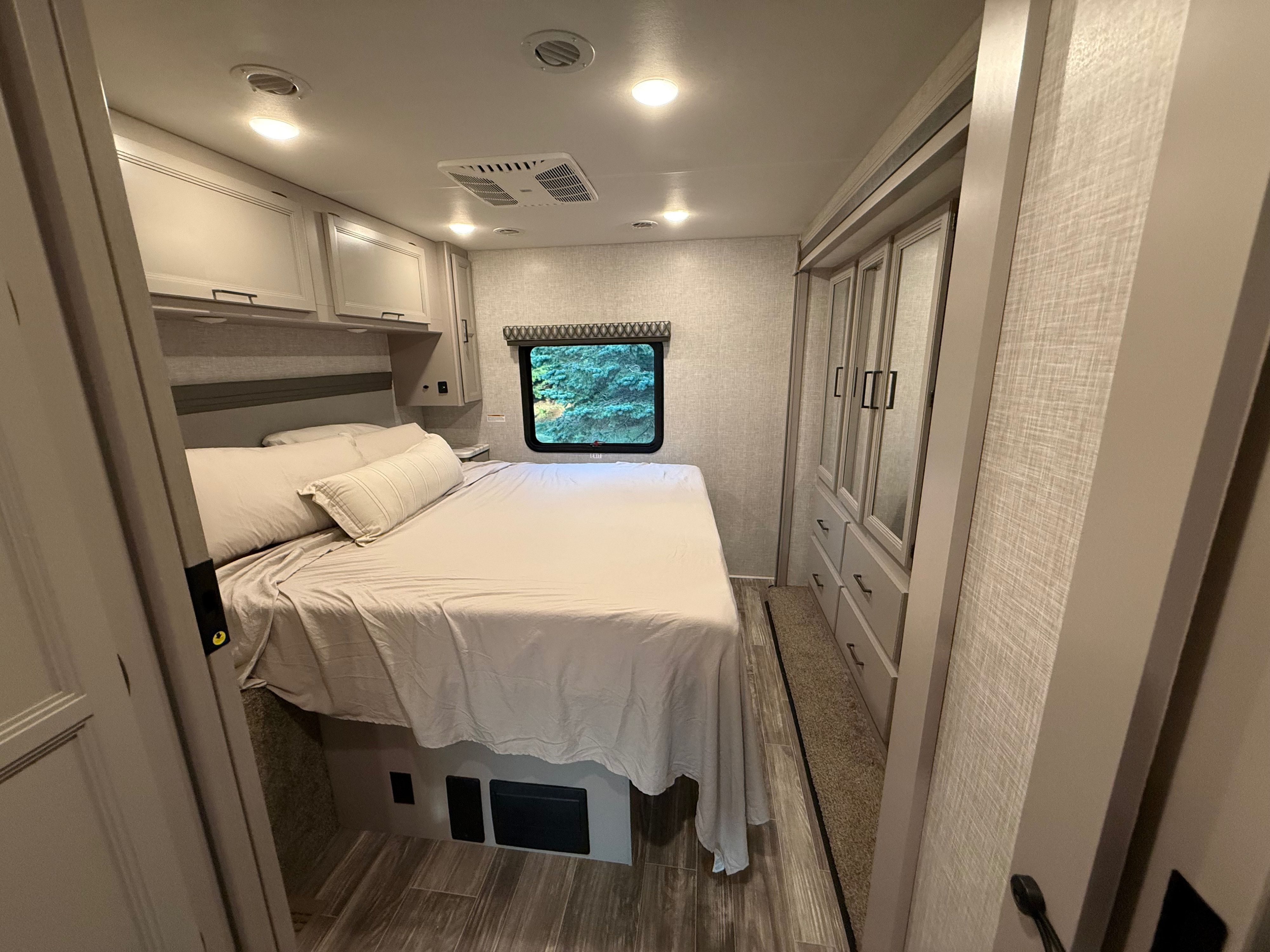 Cozy RV rental bedroom with a comfortable bed, overhead cabinets, wardrobe storage, and a window view, offering modern amenities.. Thor Hurricane 2022