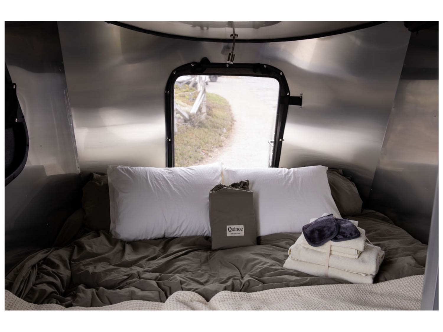 Cozy RV rental interior: bed with fresh bedding, eye mask, and a window view of an outdoor path. Relaxing travel comfort.. Airstream Basecamp 2018
