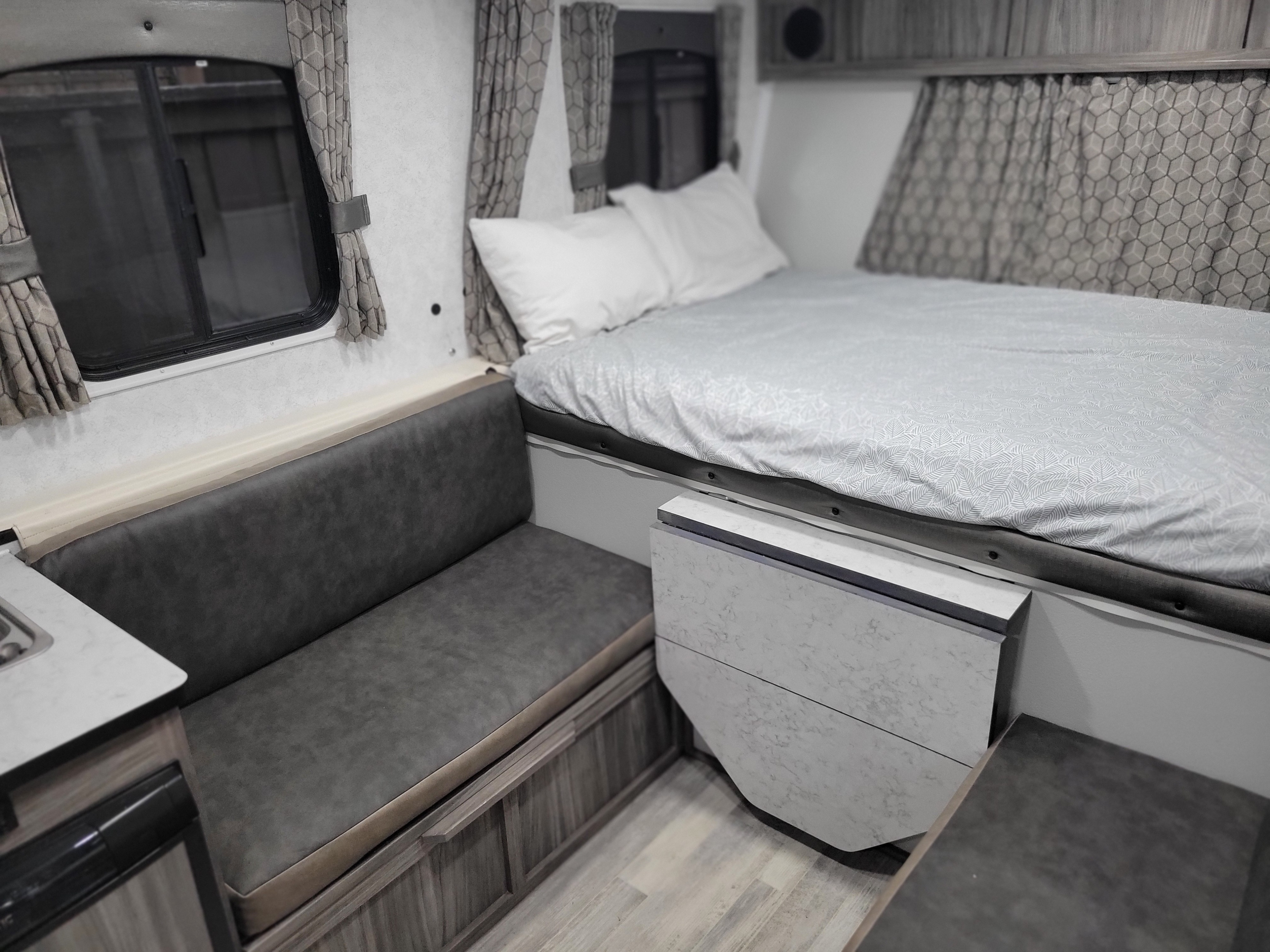 This RV rental interior features a comfortable bed, a cozy dark grey sofa, and patterned curtains by the window.. TrailManor 2518KB 2022