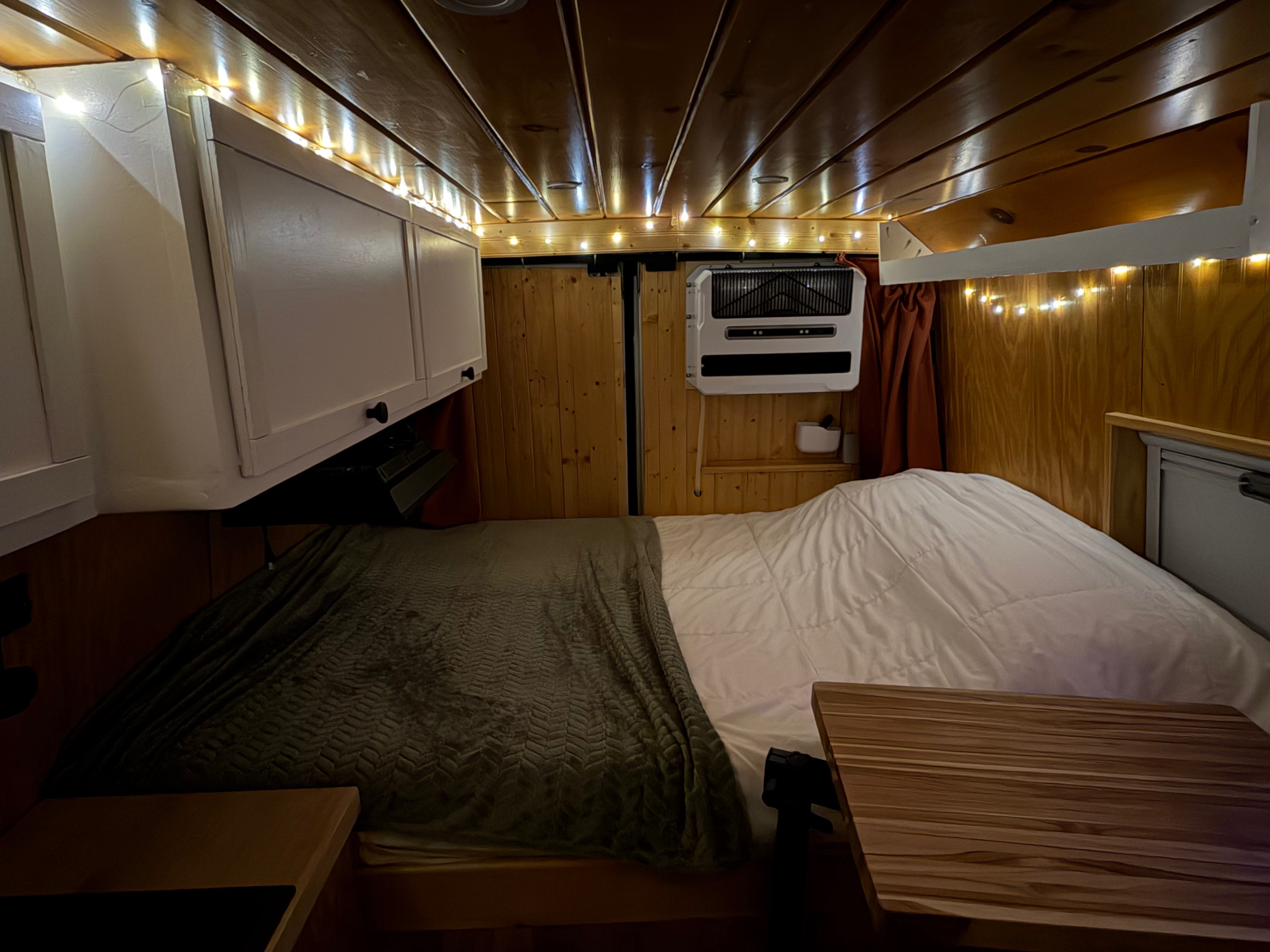 Cozy RV rental interior shows a bed, wooden walls, string lights, a/c unit, and overhead cabinets for a comfortable adventure.. Dodge Ram 2500 2024