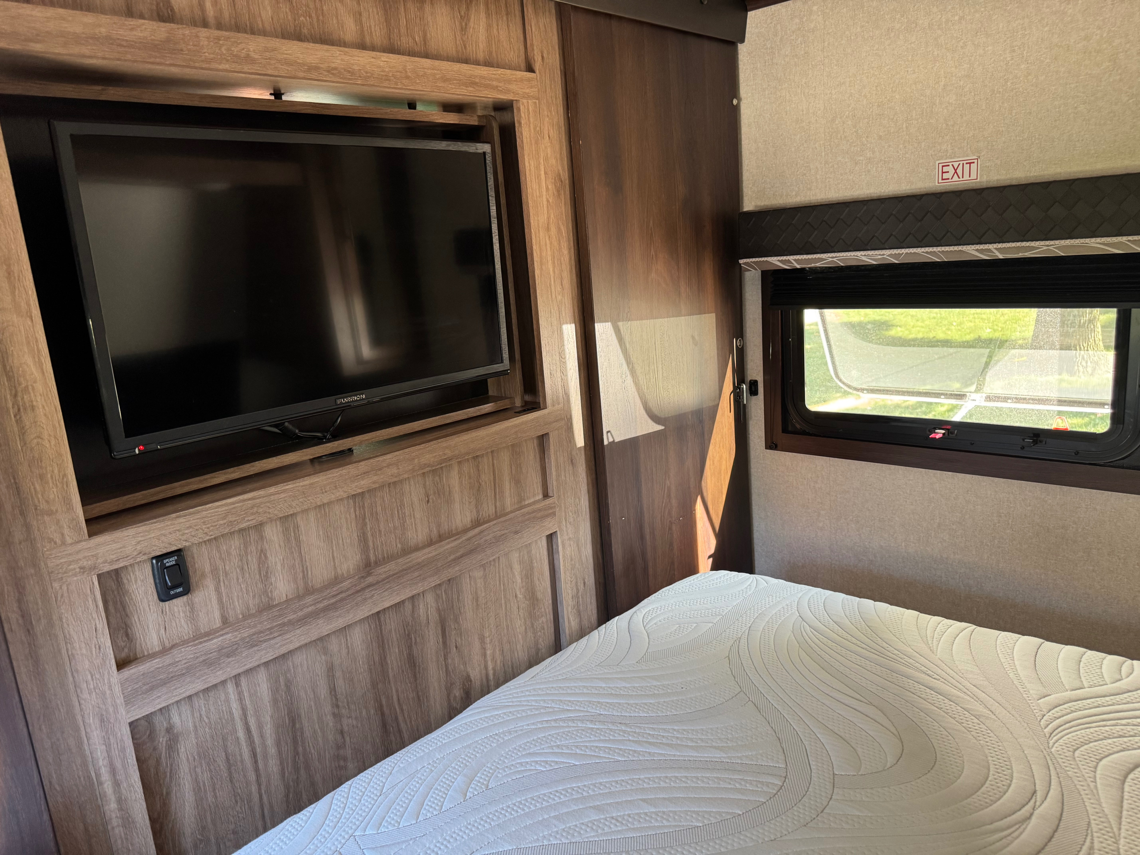 RV rental bedroom view: TV, white patterned mattress, and window with an 'EXIT' sign. Cozy wood paneling adds warmth.. Jayco Jay Feather 2018
