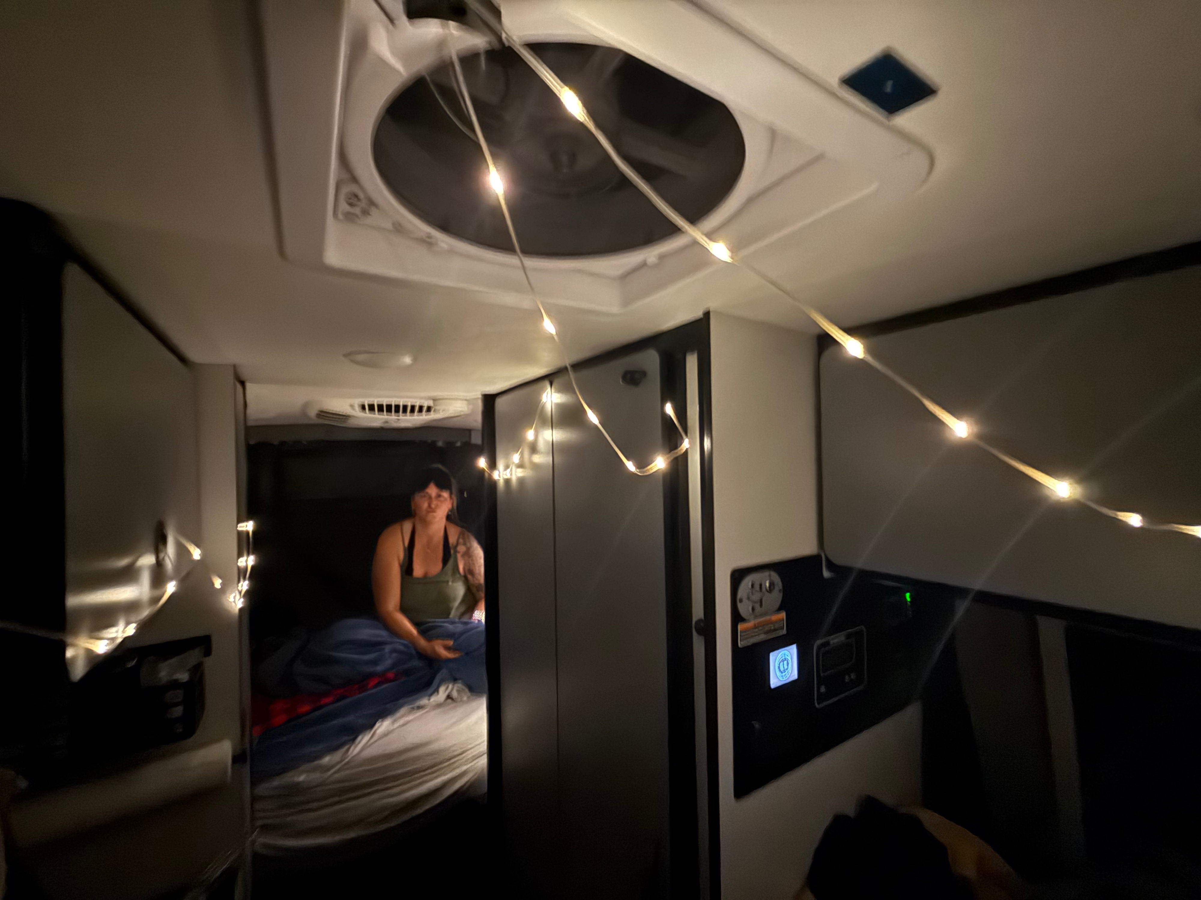 Cozy rv rental interior: a woman rests on a bed, lit by string lights. Ceiling fan & control panel visible for comfort.. Winnebago Revel 2025