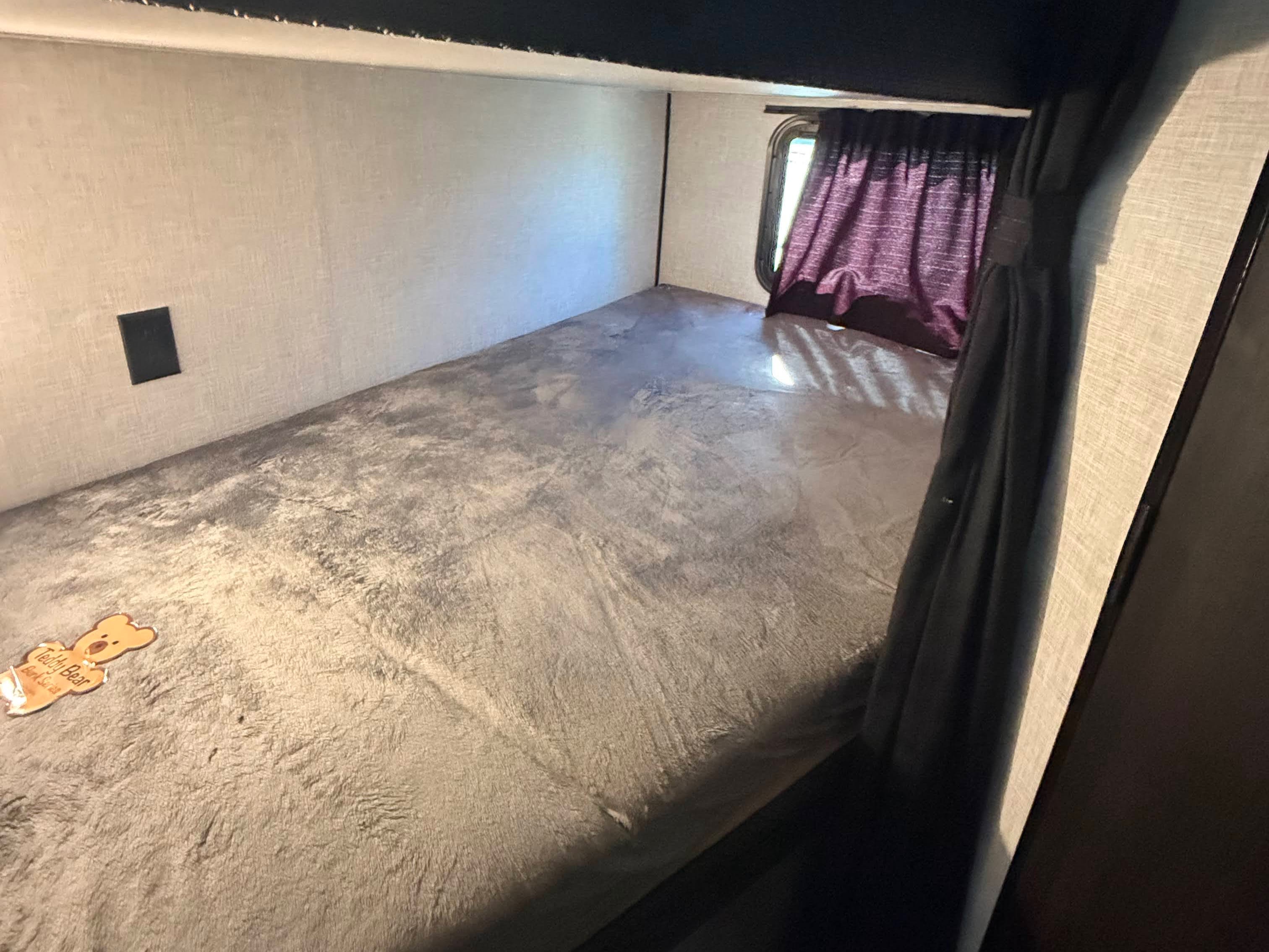 Inside an RV rental: a cozy lower bunk bed with soft grey bedding, a small window with purple curtains, and a wall outlet.. Jayco Jay Flight 2024