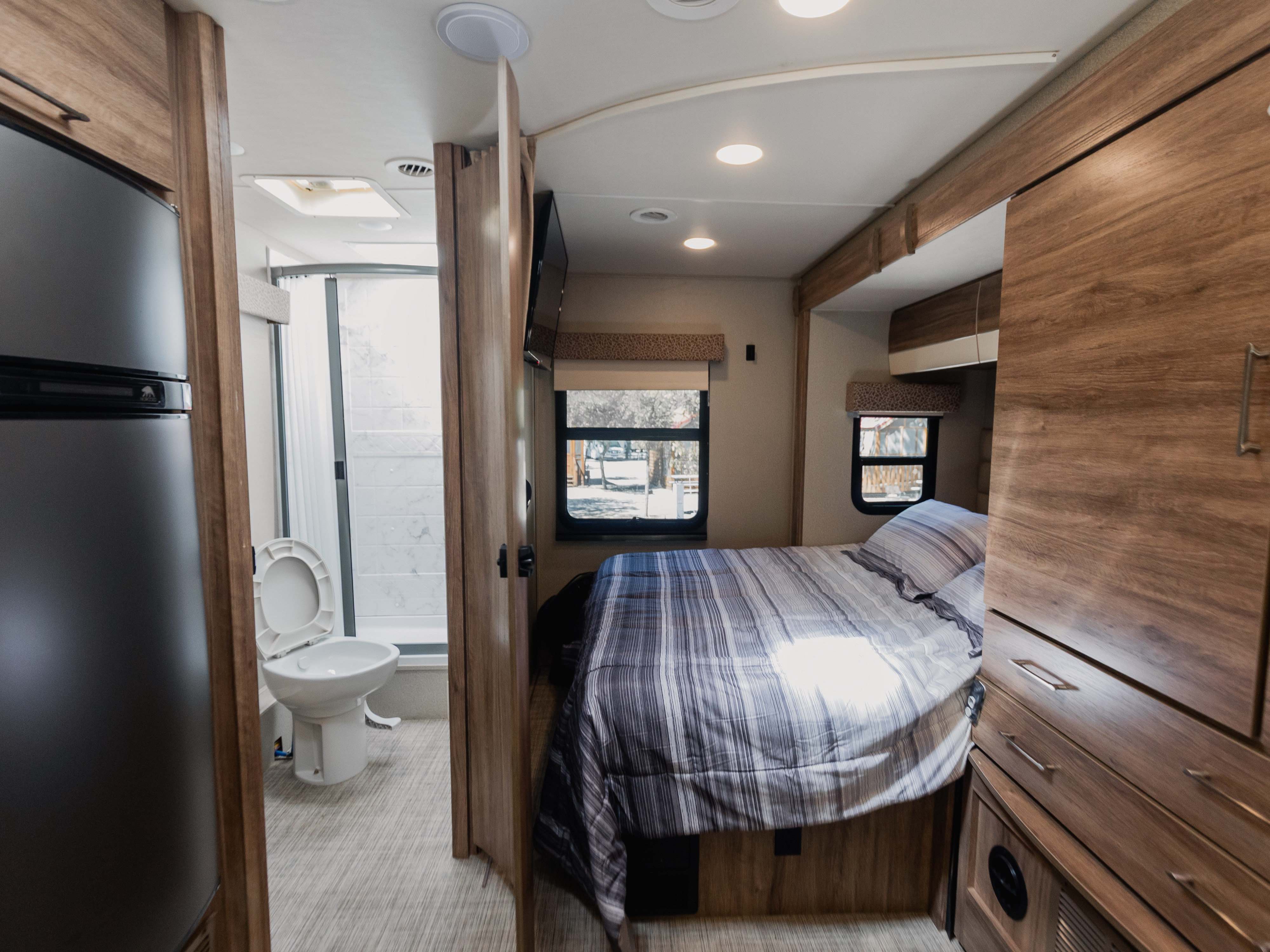 View inside an RV rental, showcasing its bedroom with patterned bedding, adjacent compact bathroom with toilet & shower, plus a fridge.. Jayco Melbourne Prestige 2020