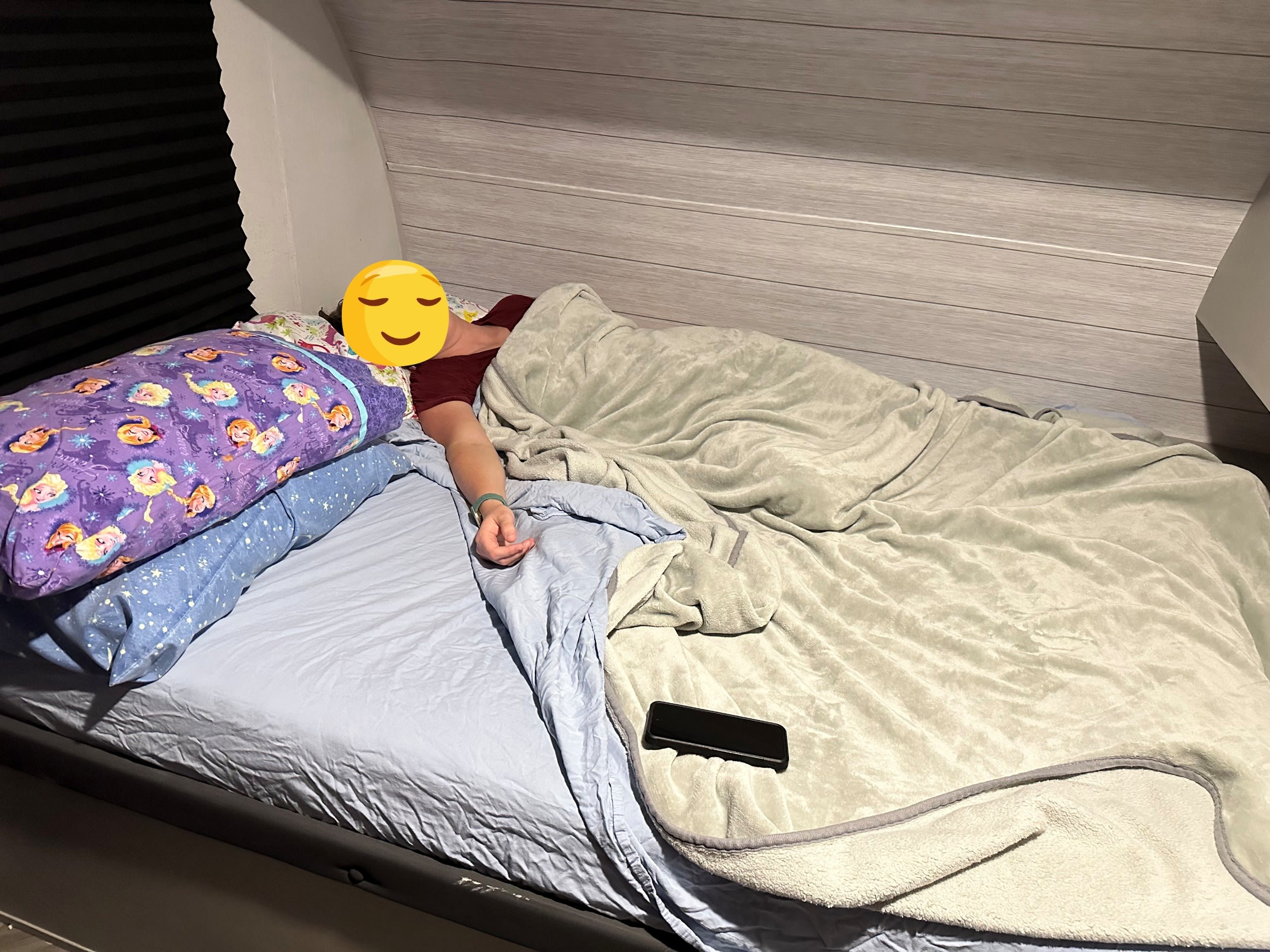 A person rests in a comfortable RV rental bed, covered by a soft blanket, with two patterned pillows and a phone nearby.. Forest River Salem FSX PLATINUM 2021
