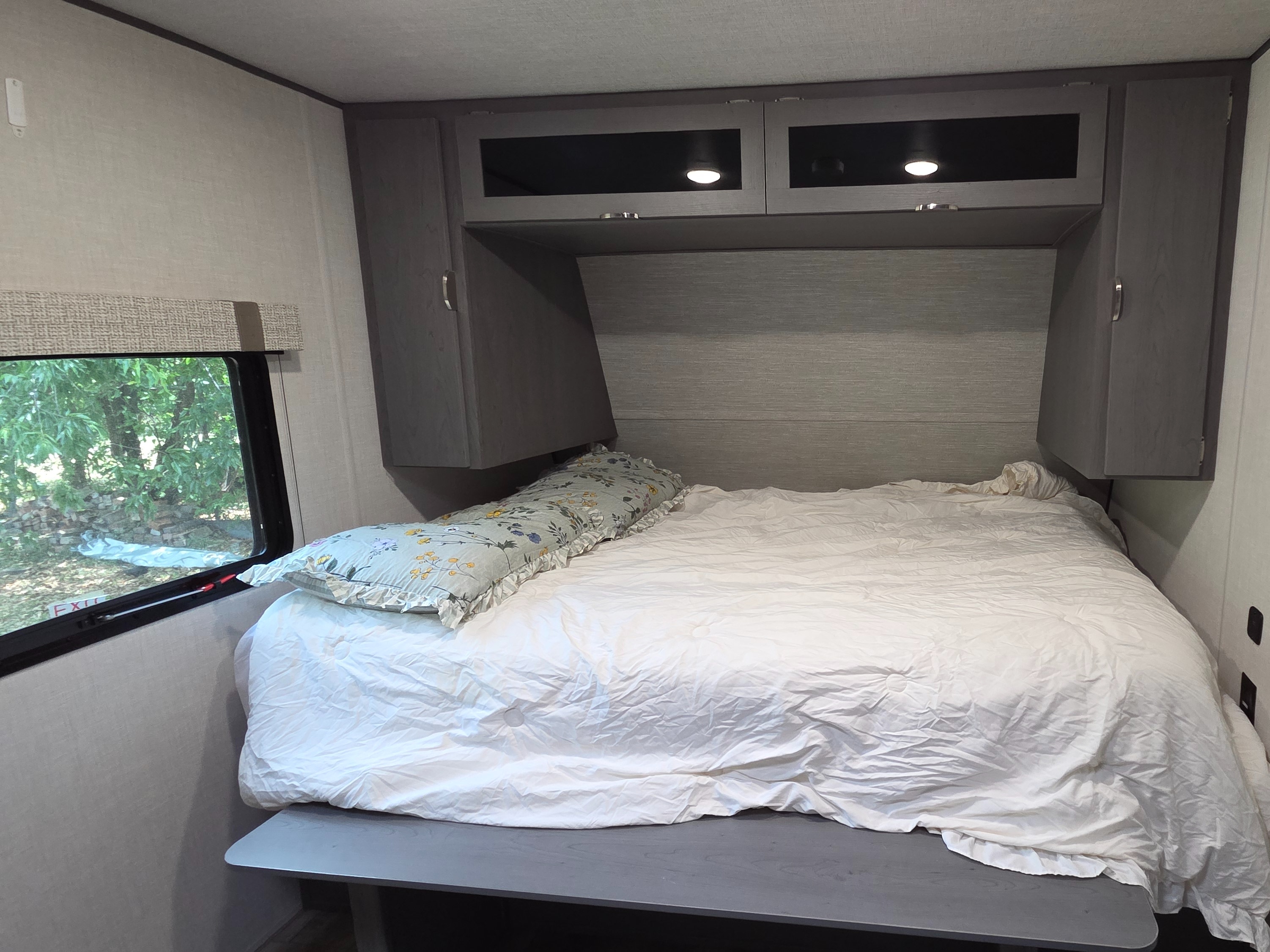 Cozy RV rental bedroom featuring a bed with white bedding, a floral pillow, overhead storage, and a window.. Ozark Ozark Trailer 2025