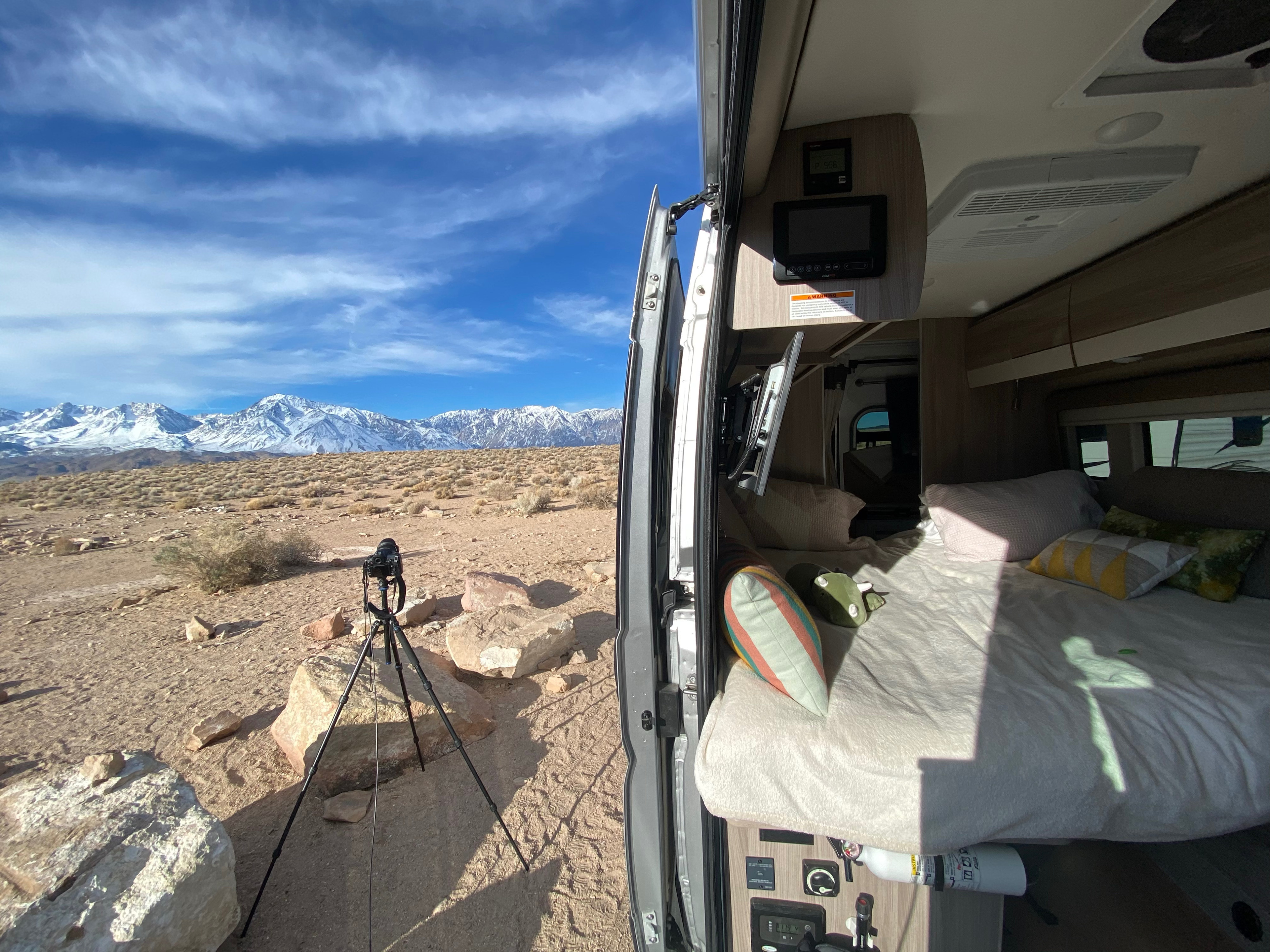RV rental van interior with bed facing majestic snow-capped mountains in a desert landscape. A camera on tripod waits.. Thor Sequence 2022