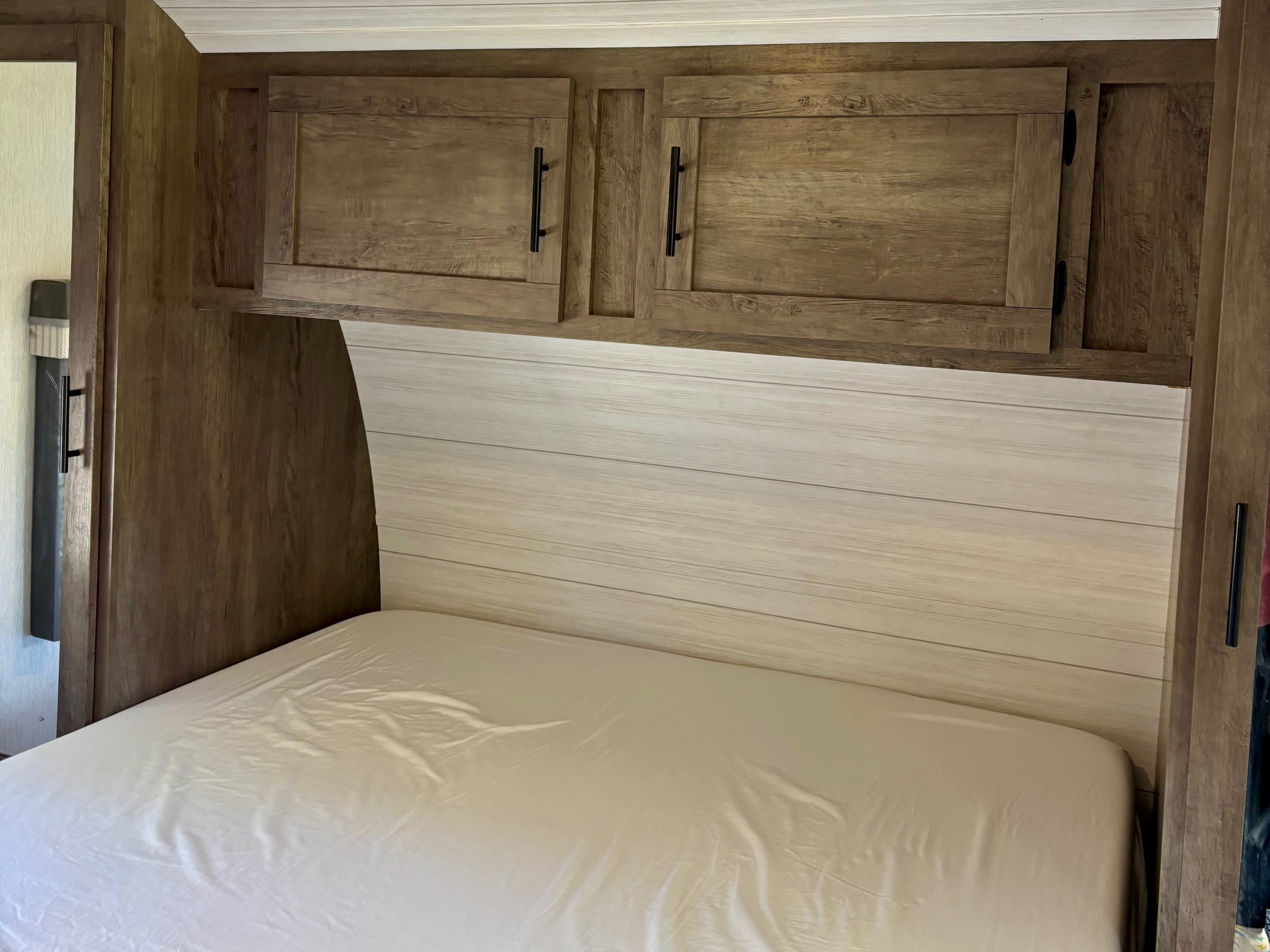 Compact RV rental bedroom featuring a neat bed, wood overhead cabinets, and a light shiplap headboard.. Forest River Salem 2022