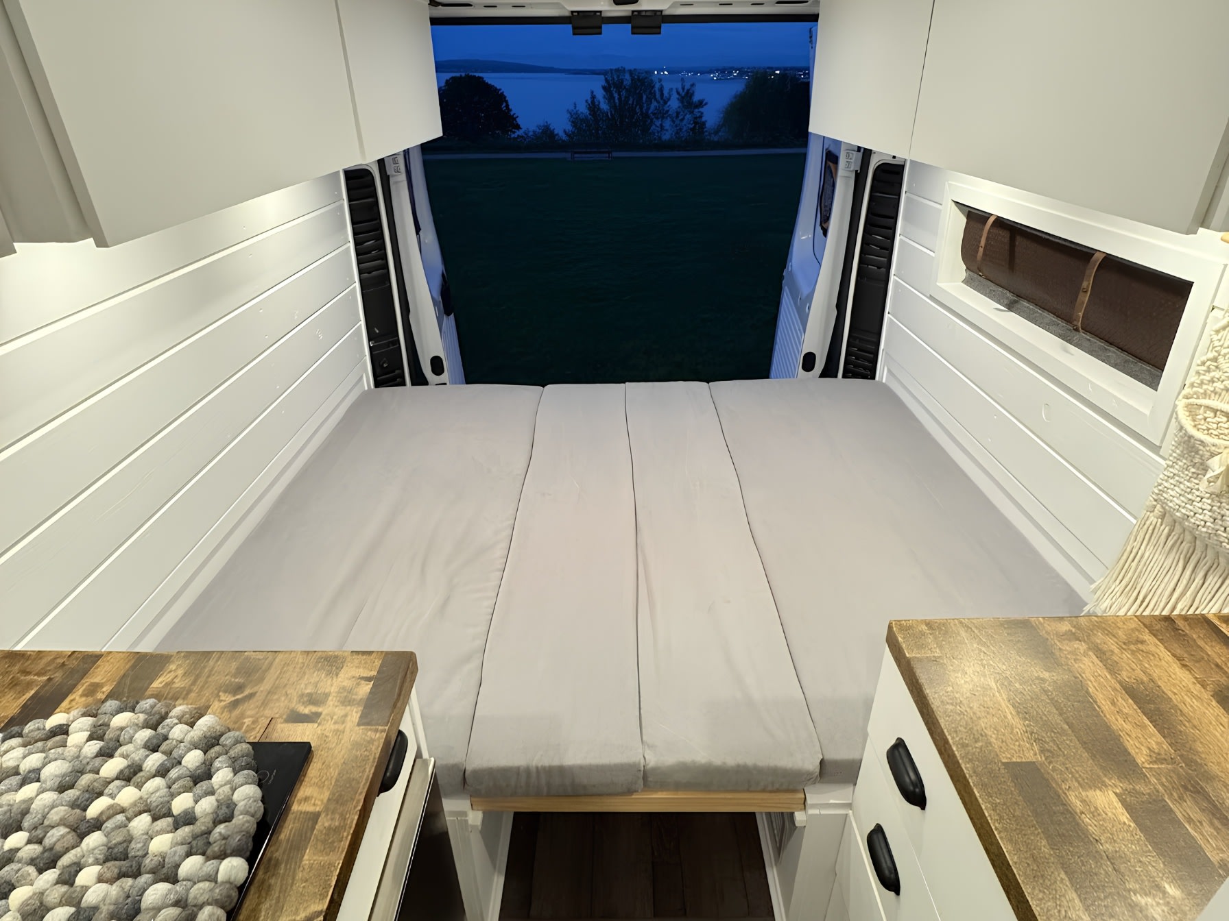 Cozy rv rental van interior with modular bed, white walls, and open rear doors revealing a tranquil twilight lake view.. RAM ProMaster 2500 2018