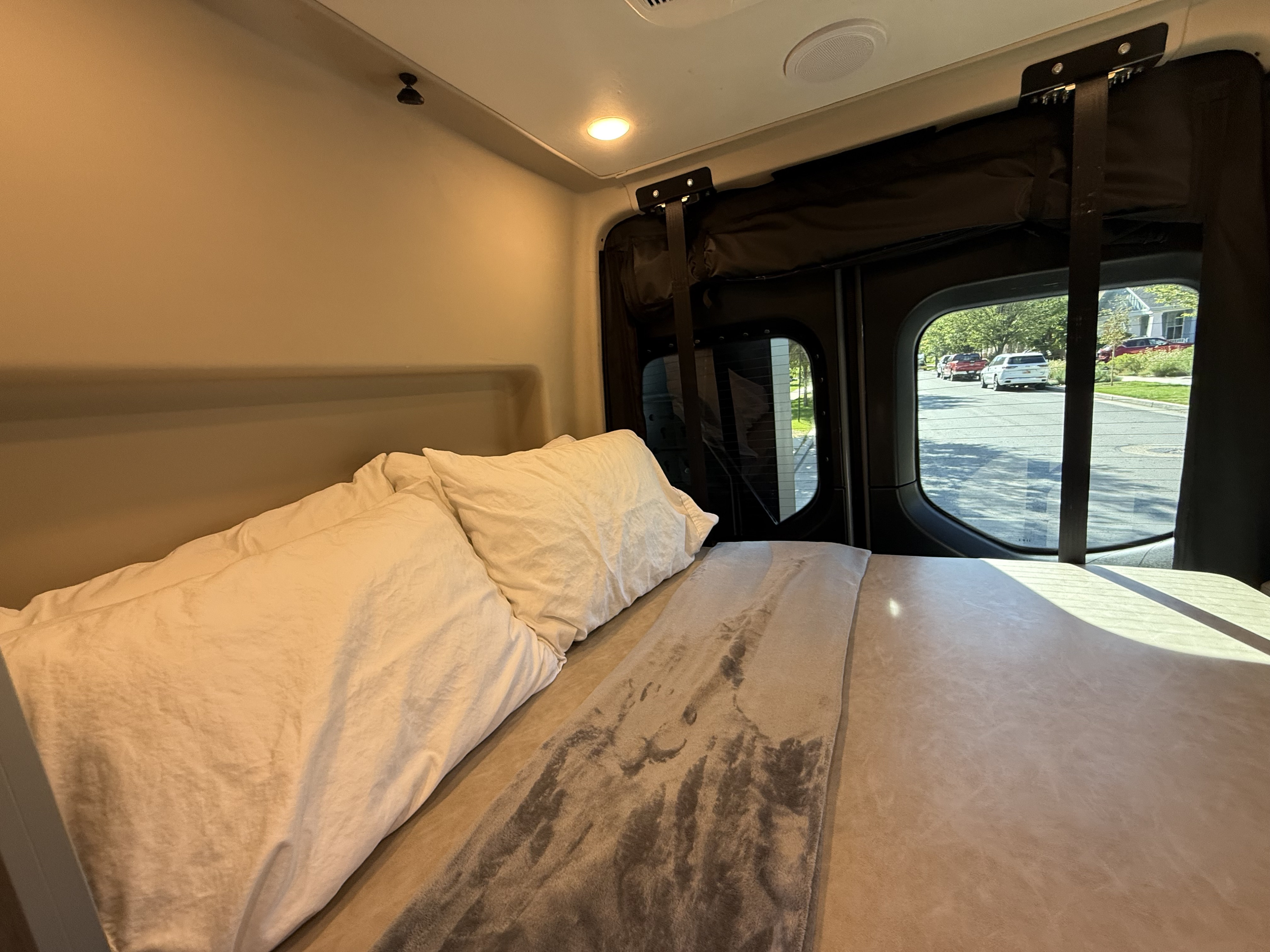 This inviting rv rental features a cozy bed with white pillows and a gray blanket, plus a street view outside the window. Ideal for travel.. Jayco Terrain 2023