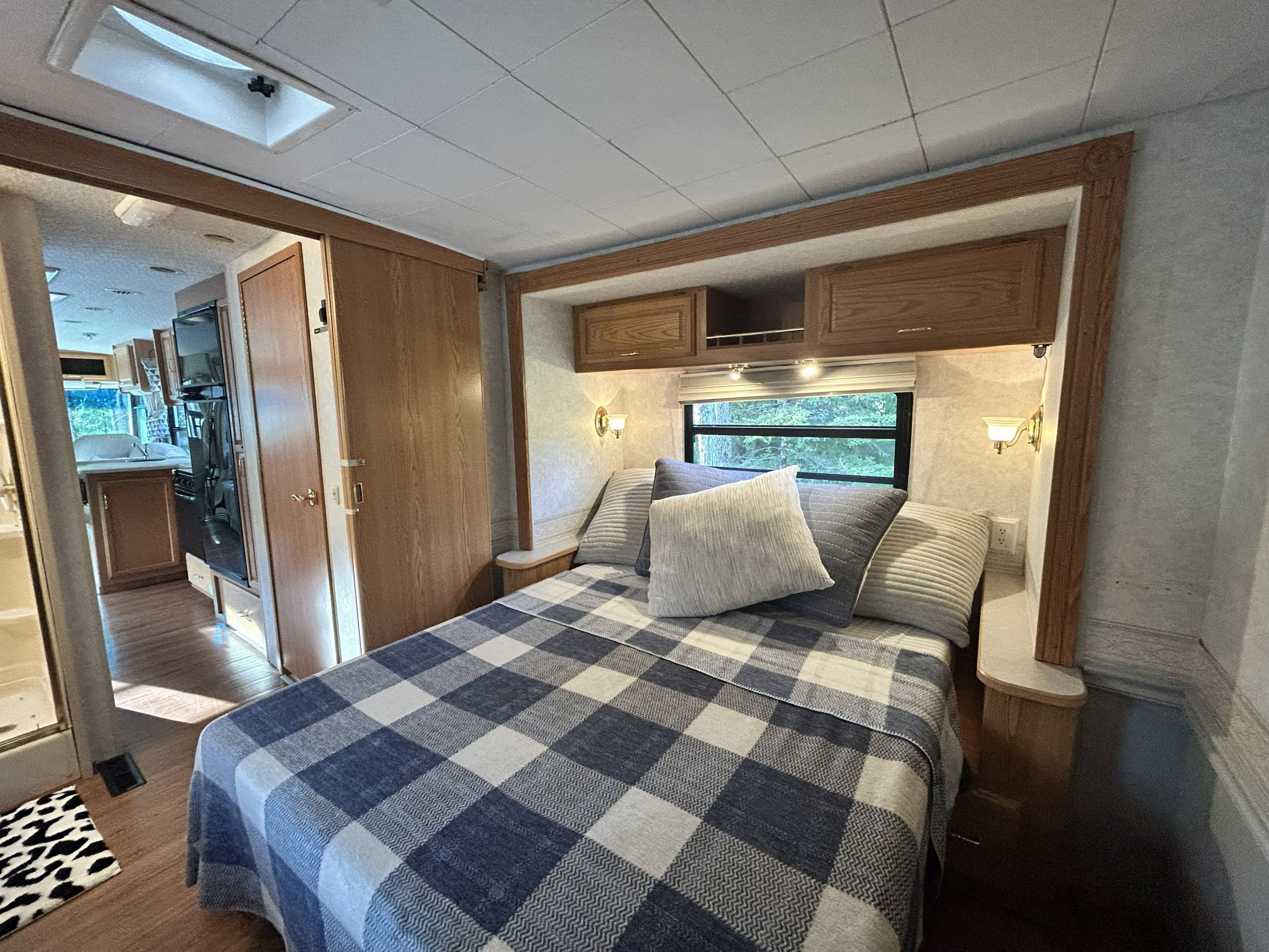 This RV rental's cozy bedroom features a plaid bed, overhead cabinets, a window with a treed view, and wood floors.. Winnebago Brave 2004