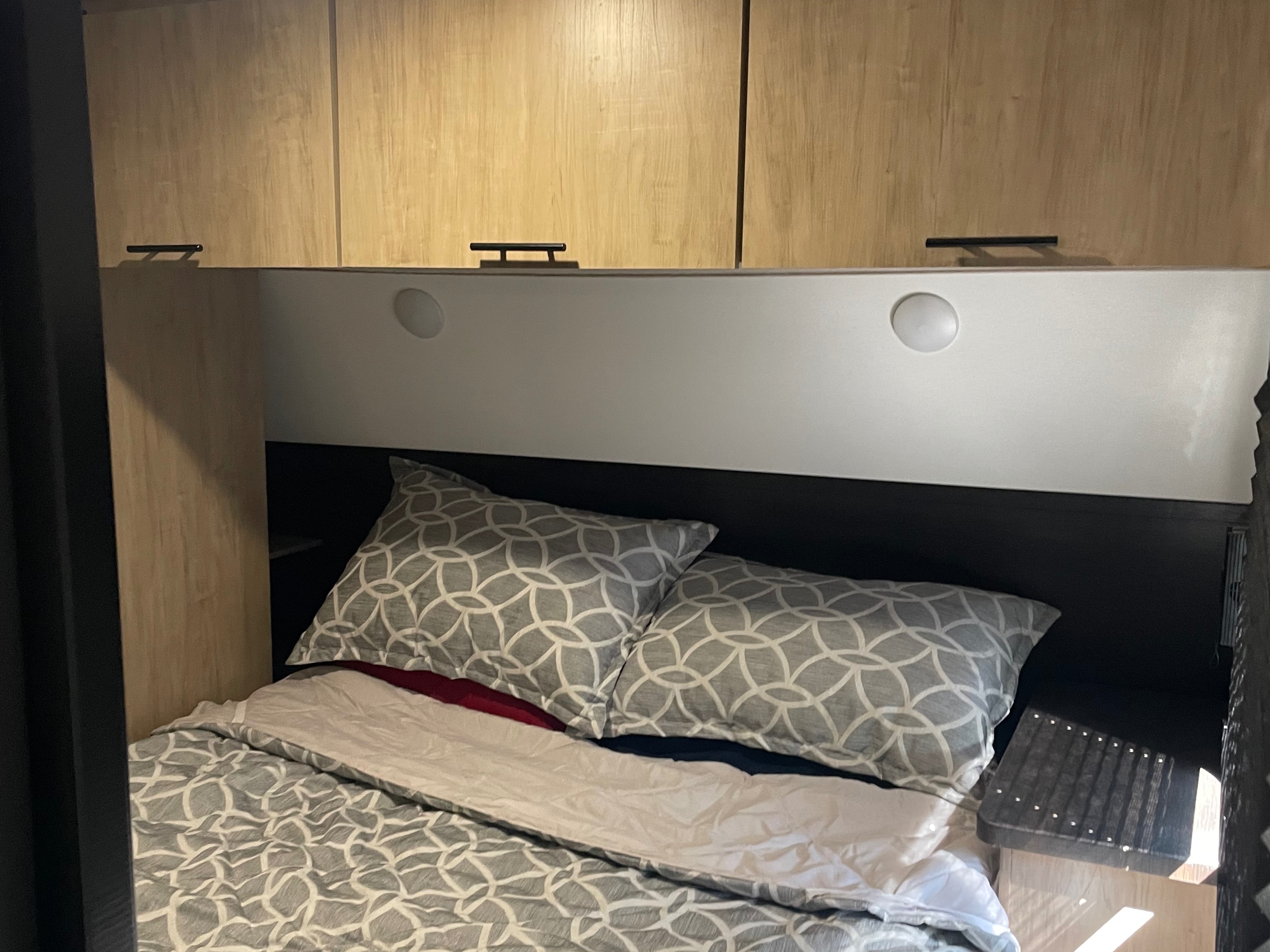 A compact RV rental bedroom with patterned bedding, a dark headboard, and light wood overhead cabinets with recessed lighting.. Forest River Cherokee Grey Wolf 2026
