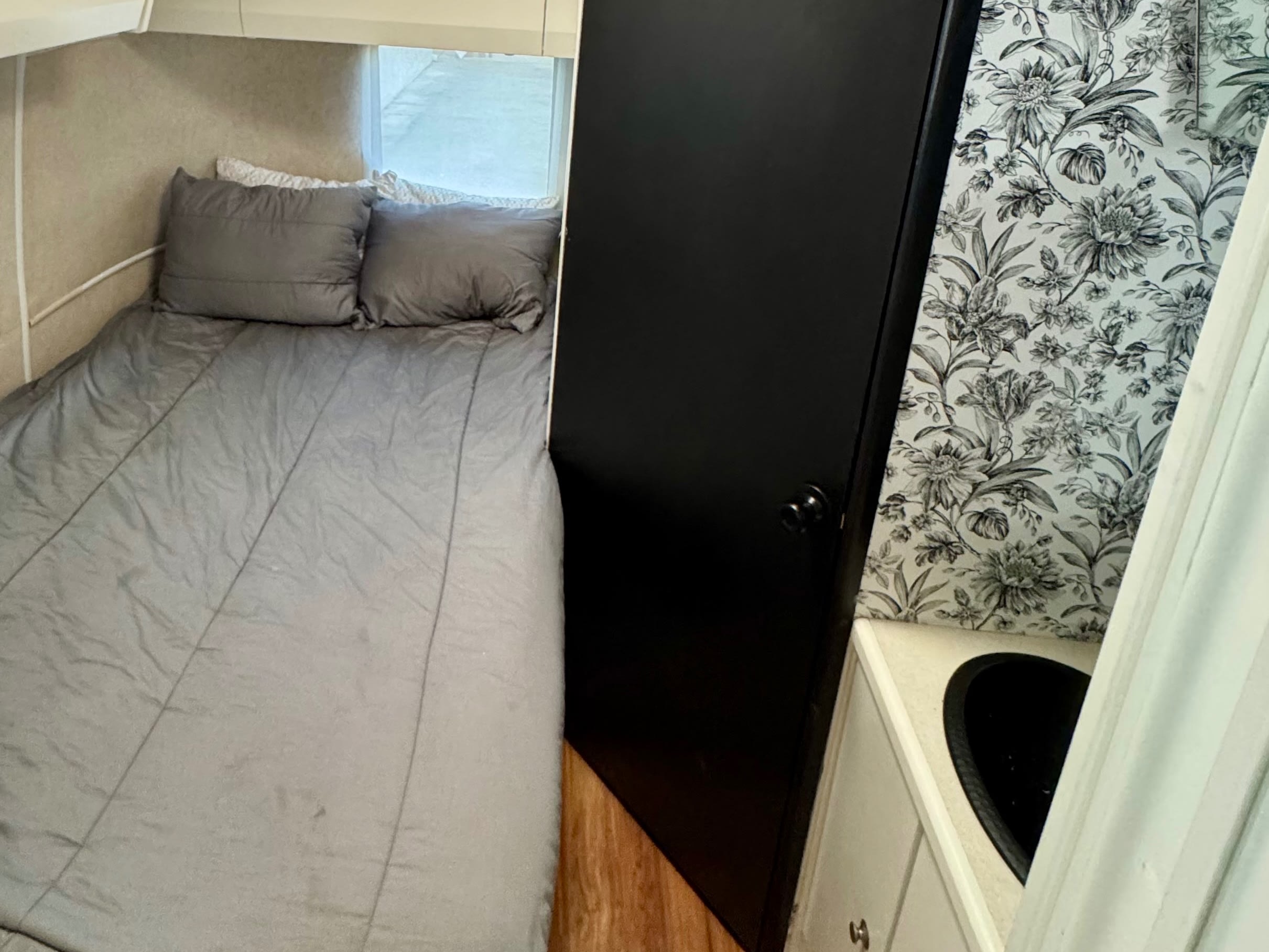 Cozy rv rental interior with a gray bed, dark door, and a compact bathroom sink area featuring floral wallpaper.. Airstream Bambi 1999