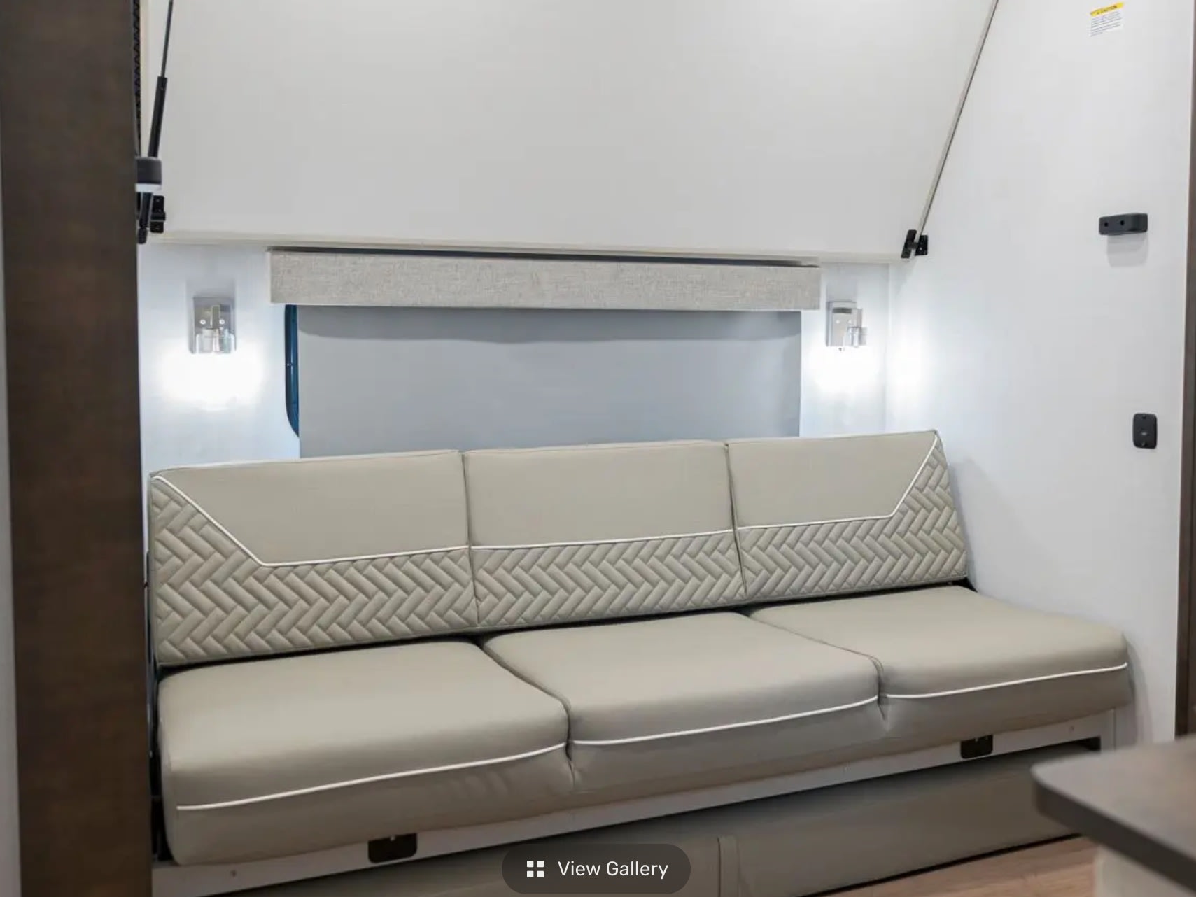 Modern RV rental interior with a light beige sofa featuring quilted backrests and white piping, flanked by bright wall lights.. Alliance RV Avenue 2026