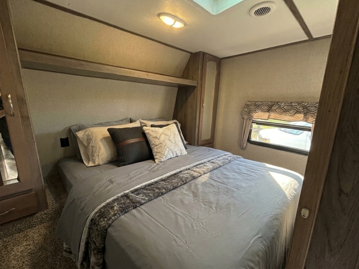 Relax in this comfortable RV rental bedroom. Features a queen bed with gray bedding, pillows, overhead shelf, and a window.. Keystone RV Sprinter Campfire Edition 2018