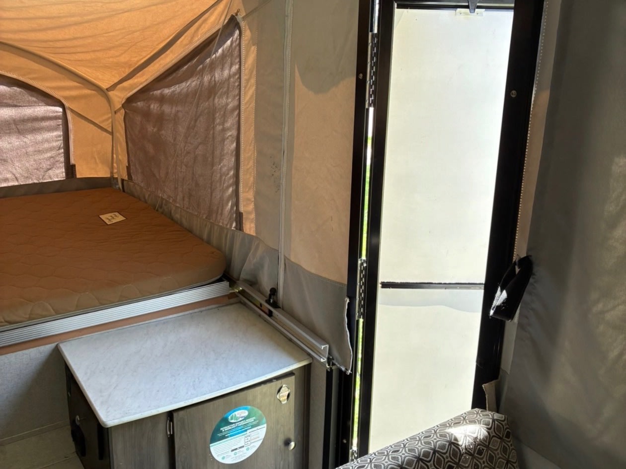 Inside this RV rental pop-up camper: a bed with a brown mattress, a small counter, and the entry door on the right.. Coachmen clipper 2020