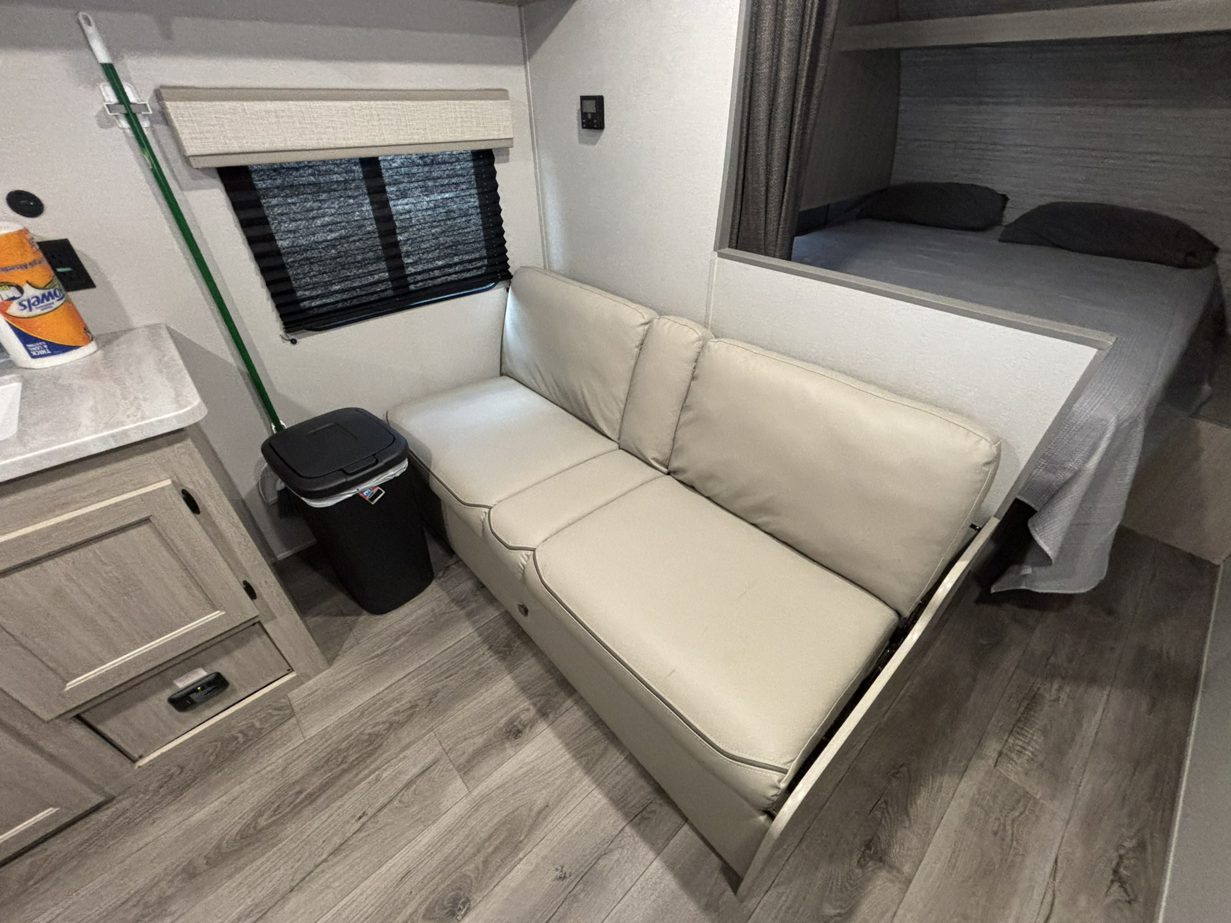 Cozy rv rental interior with a light sofa, kitchen counter, trash can, mop, and a compact bed area behind a partition.. Coachmen 261BH 2024