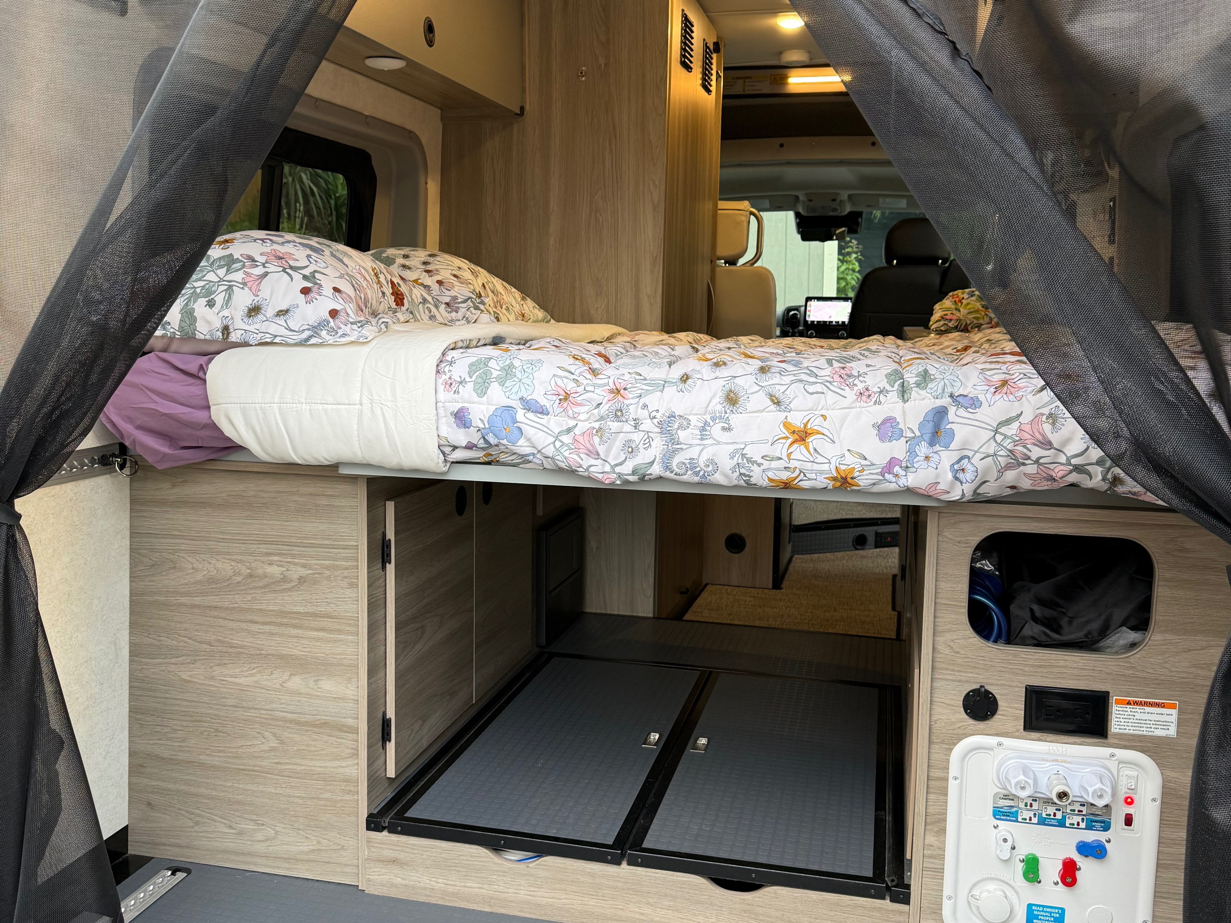 Cozy RV rental interior featuring a raised bed with floral bedding, under-bed storage, rear screen netting, and utility hookups.. Winnebago Solis 2023