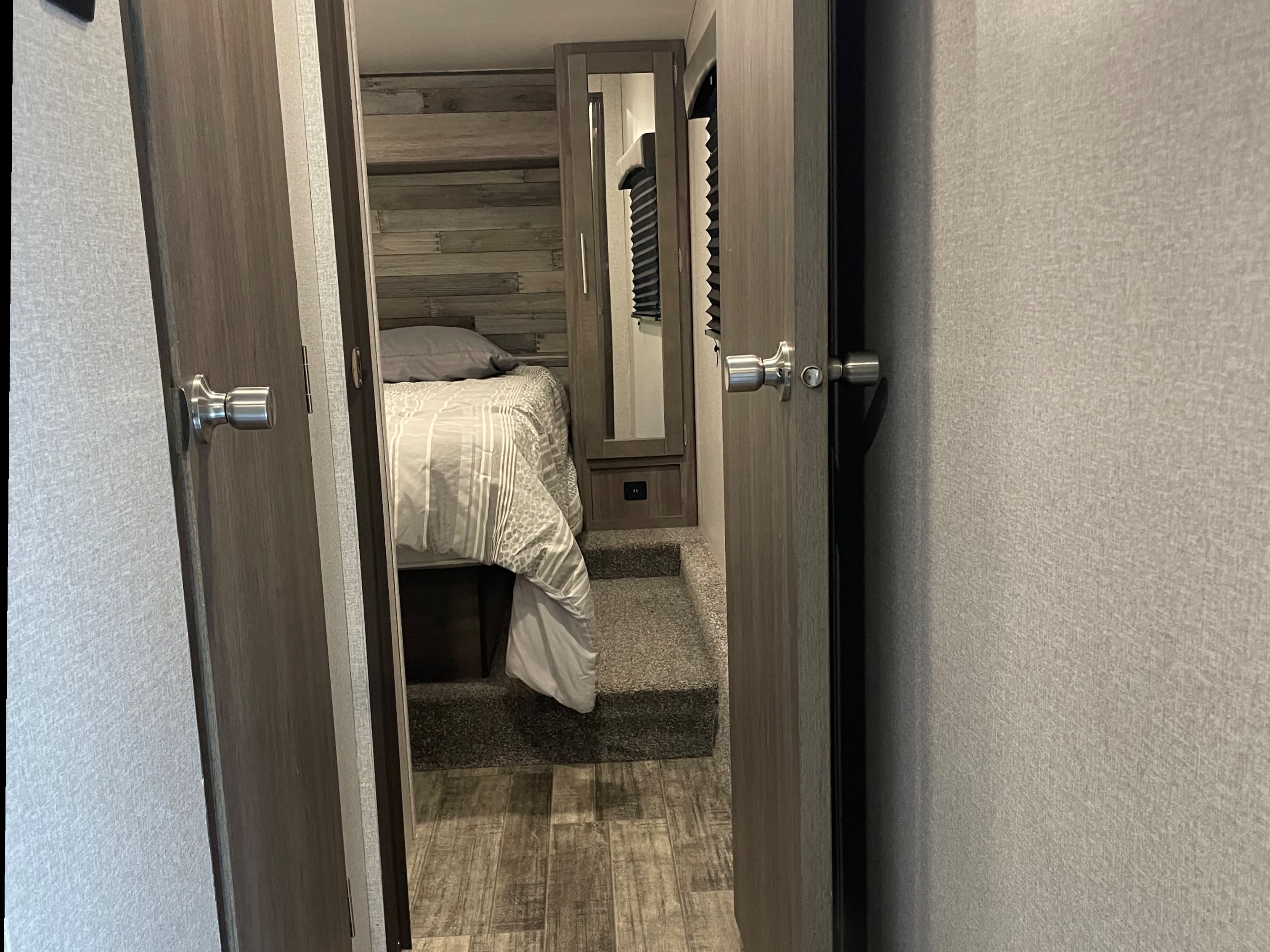 Cozy bedroom in an RV rental, seen through open doors. Features a bed, wood accents, and carpeted steps.. Crossroads RV Volante 2021