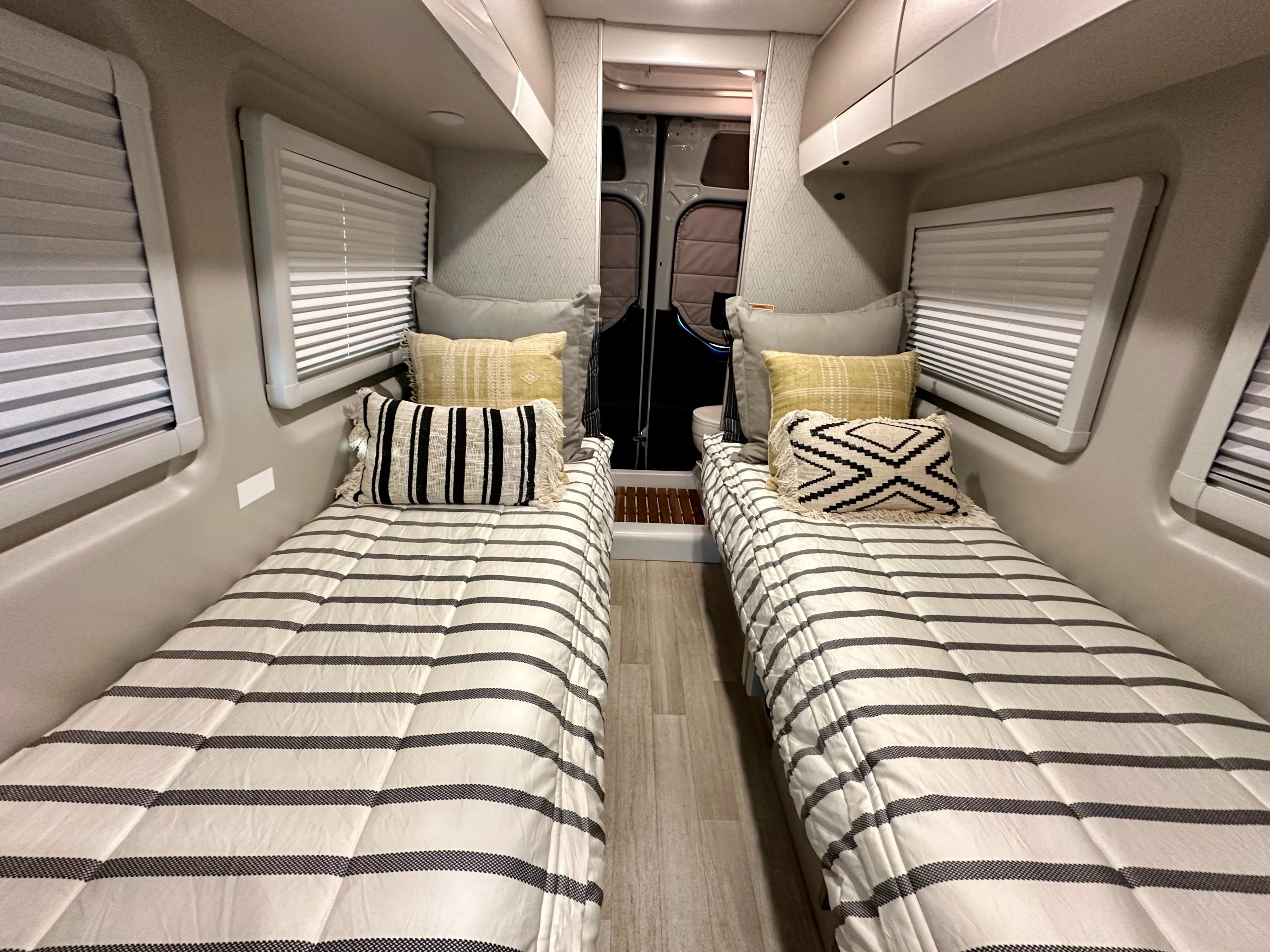 Comfortable RV rental interior with two cozy twin beds, striped comforters, decorative pillows, and window blinds for privacy.. Thor Tranquility 2026