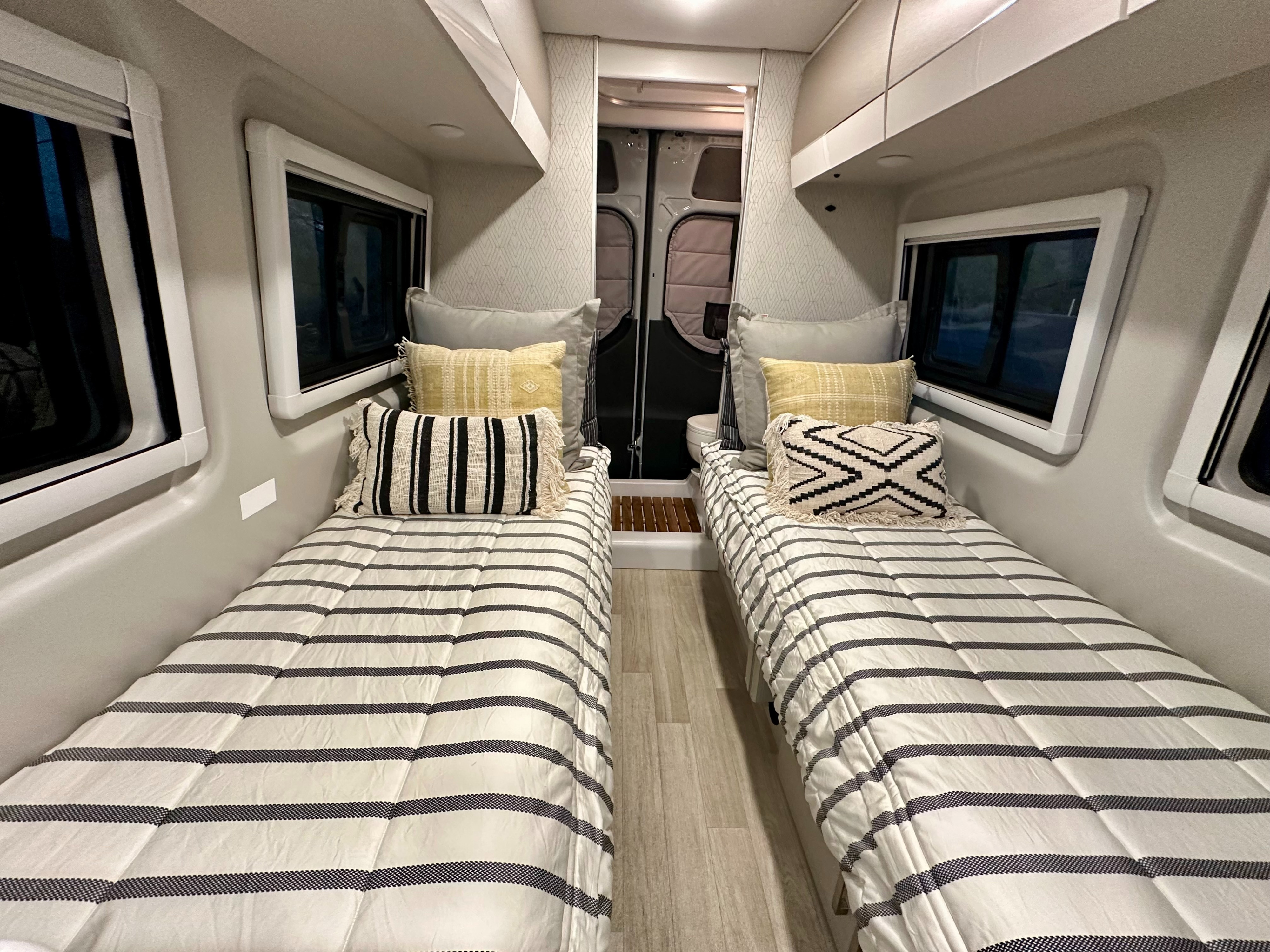 Inside an RV rental, two beds with striped comforters and accent pillows provide a cozy sleeping area.. Thor Tranquility 2026