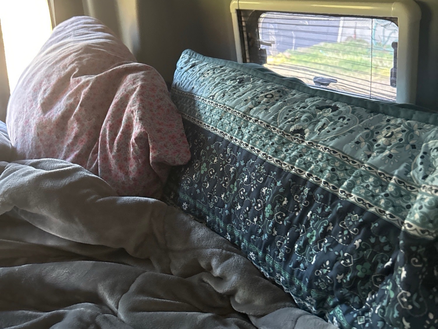 Cozy RV rental interior with person sleeping under floral blanket, window view, and patterned pillow.. Mercedes-Benz Revel 2022