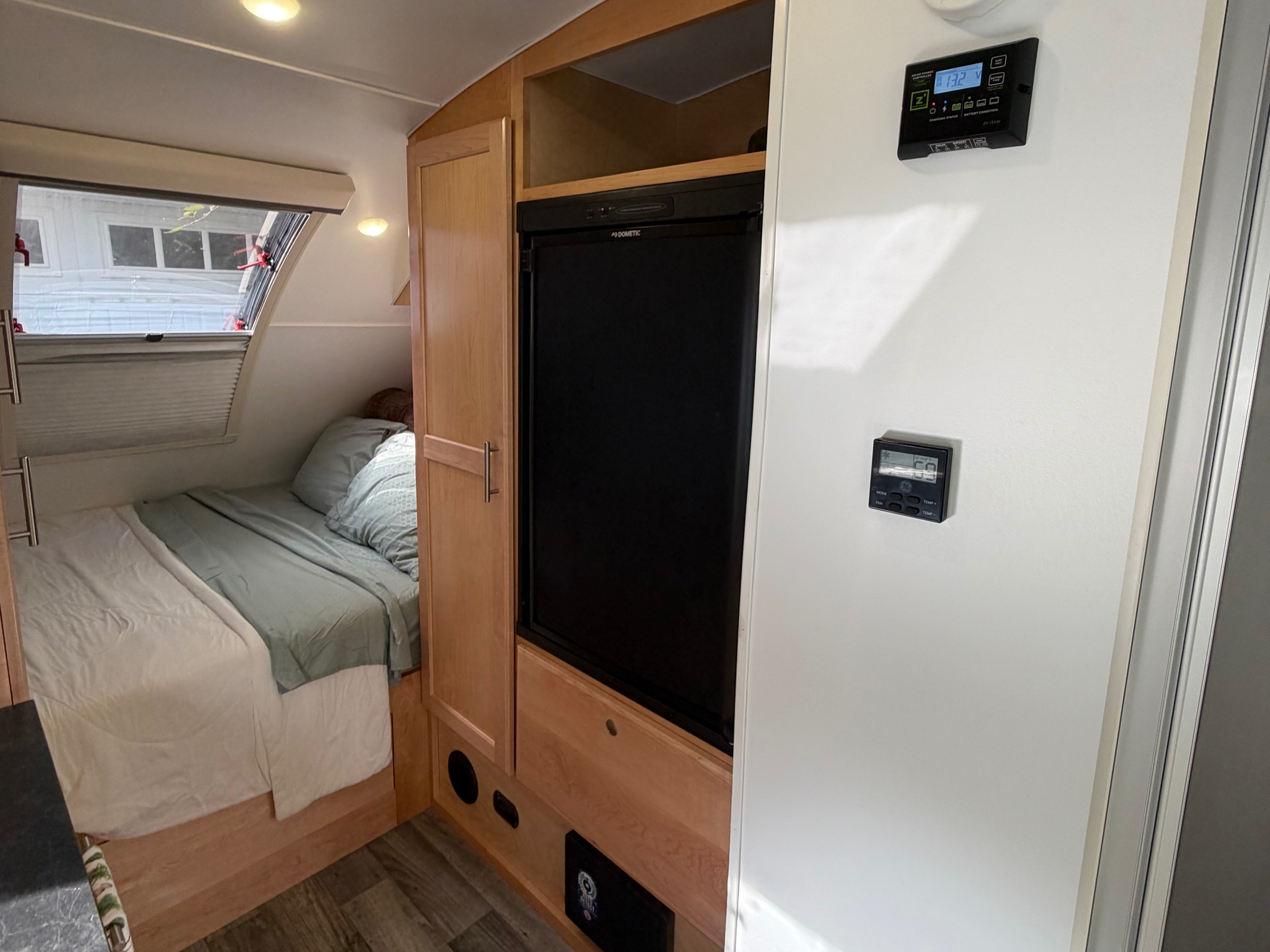Inside a comfortable RV rental: a compact bedroom with a window, fridge, and climate controls on the wall.. Little Guy Little Guy Trailer 2023