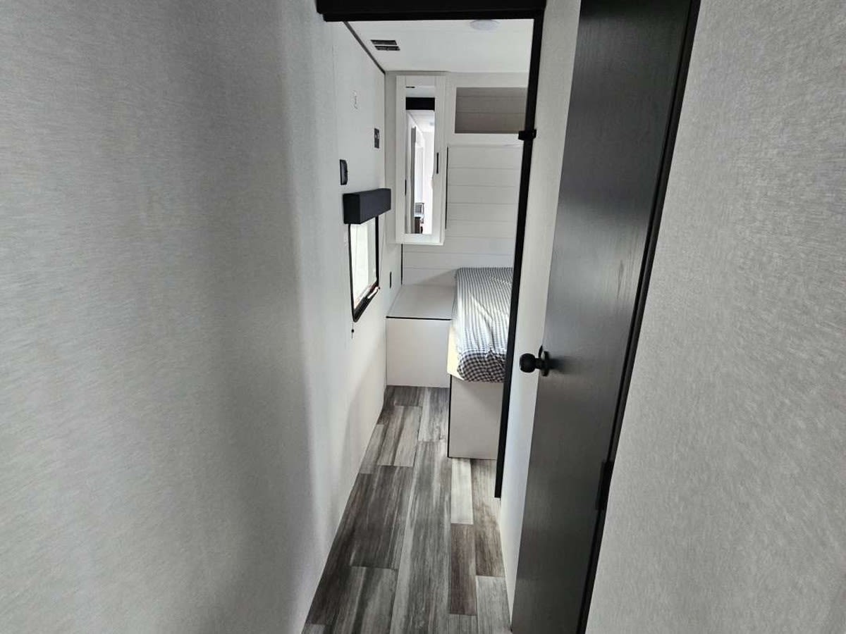 This RV rental shows a narrow, modern hallway leading to a compact bedroom with a striped bed and light walls.. Jayco Jay Flight SLX 2026