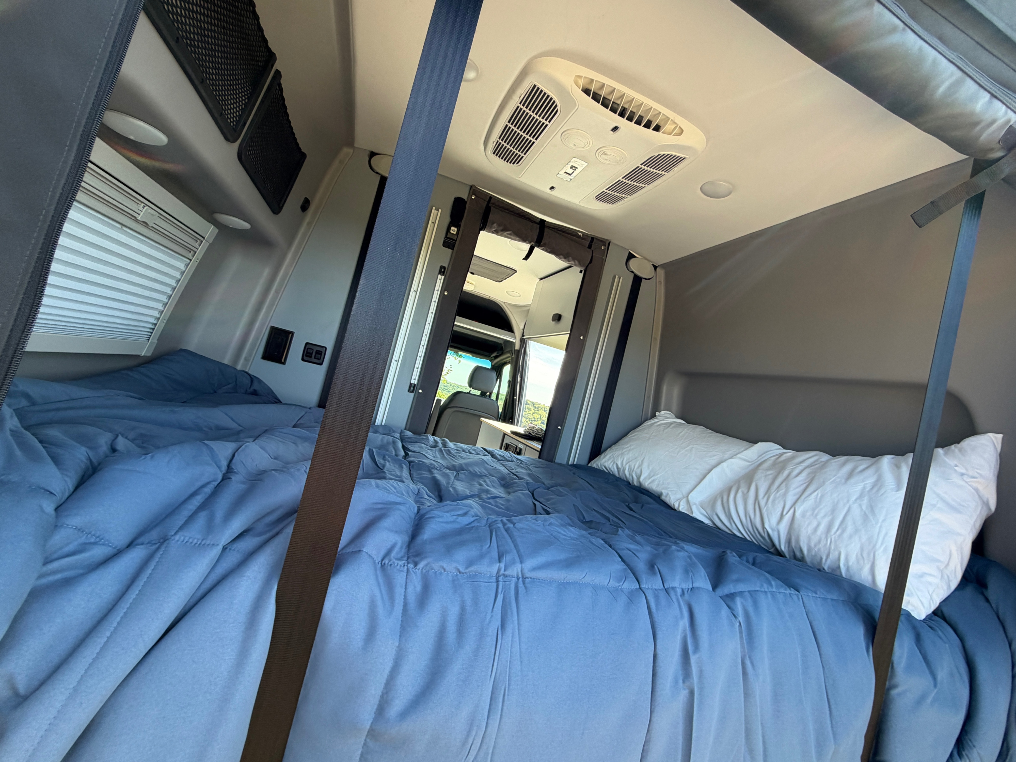 Cozy RV rental interior: comfortable bed with blue comforter, white pillow, and overhead AC unit in a camper van.. Winnebago Revel 2025
