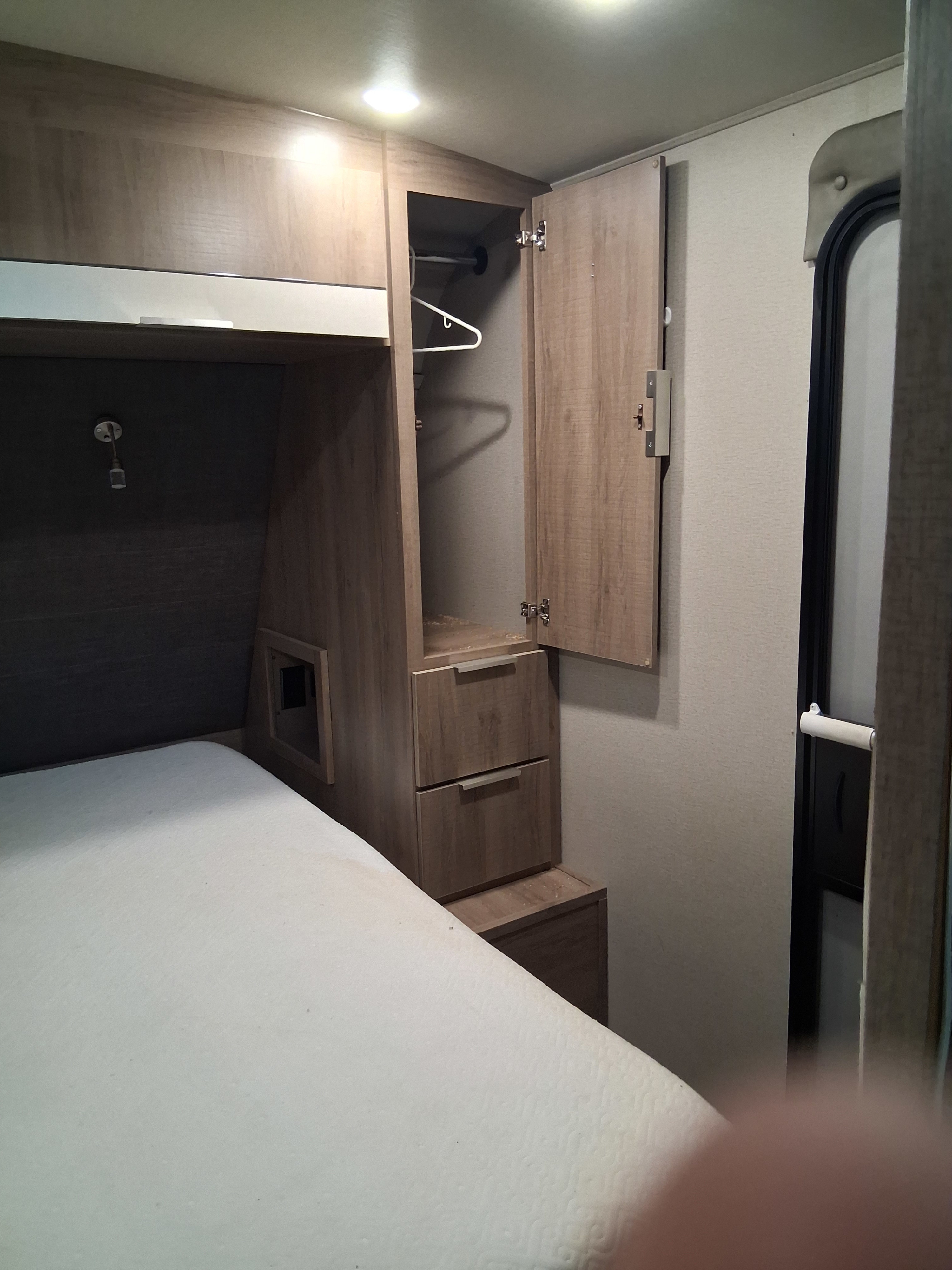 RV rental bedroom view: comfortable bed, open wood wardrobe with hanger, drawers, overhead cabinets, and an entry door.. Grand Design Imagine 2021