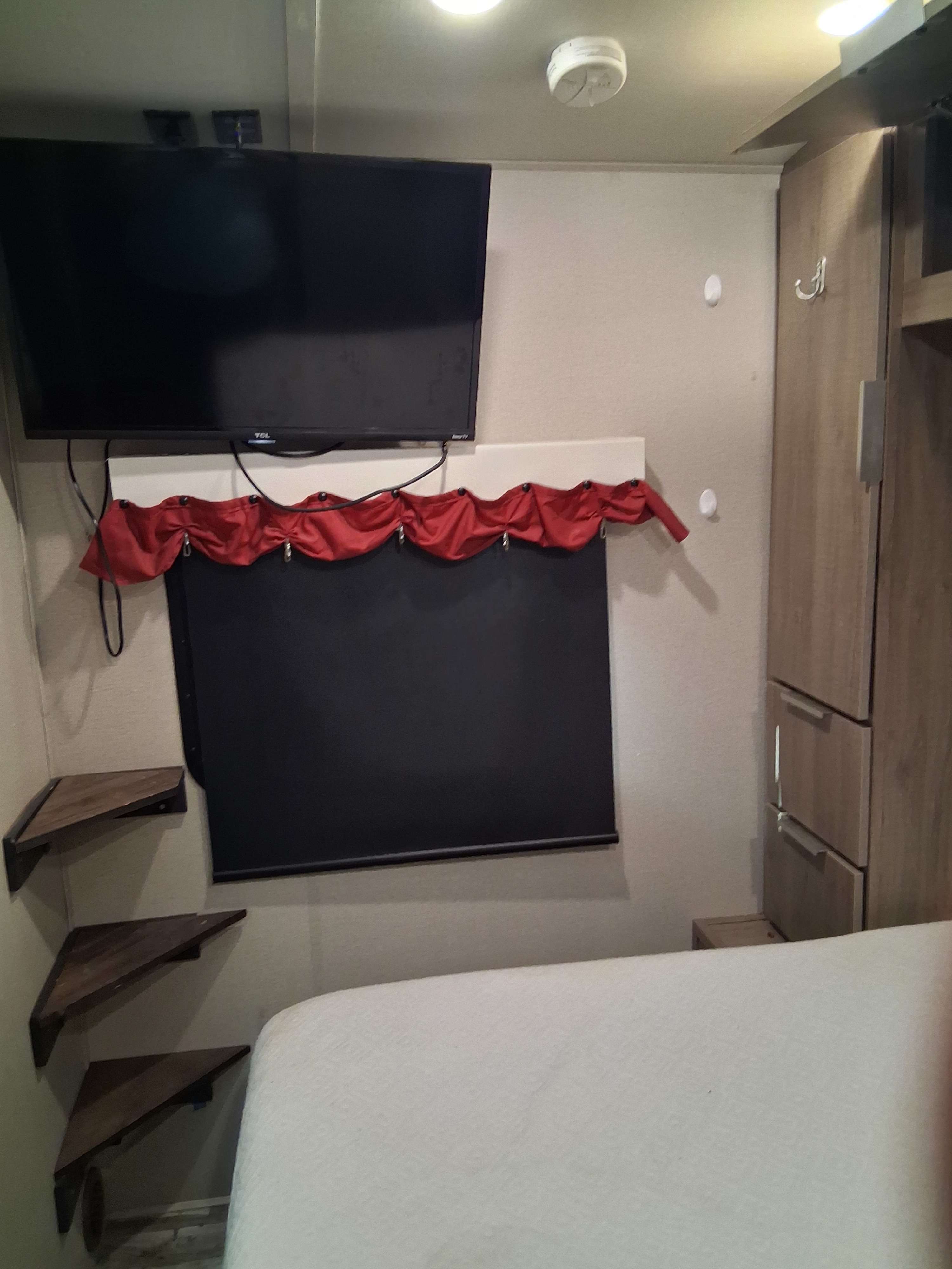 Compact RV rental bedroom featuring a wall-mounted TV, window blind, corner shelves, and a comfortable bed.. Grand Design Imagine 2021