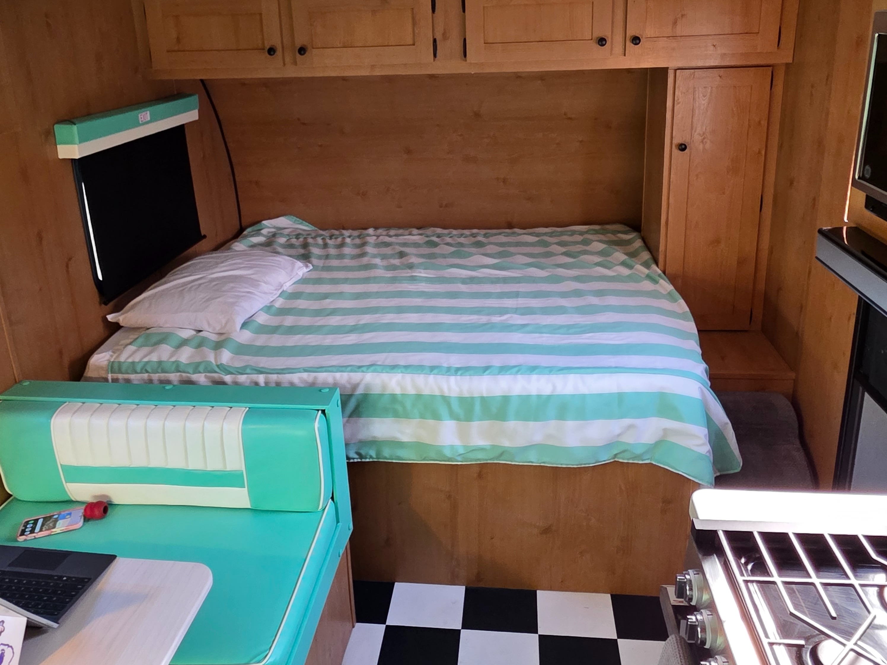 This rv rental interior features a bed with teal striped bedding, wooden walls, a teal dinette, and a checkered floor.. Riverside RV Retro 2024