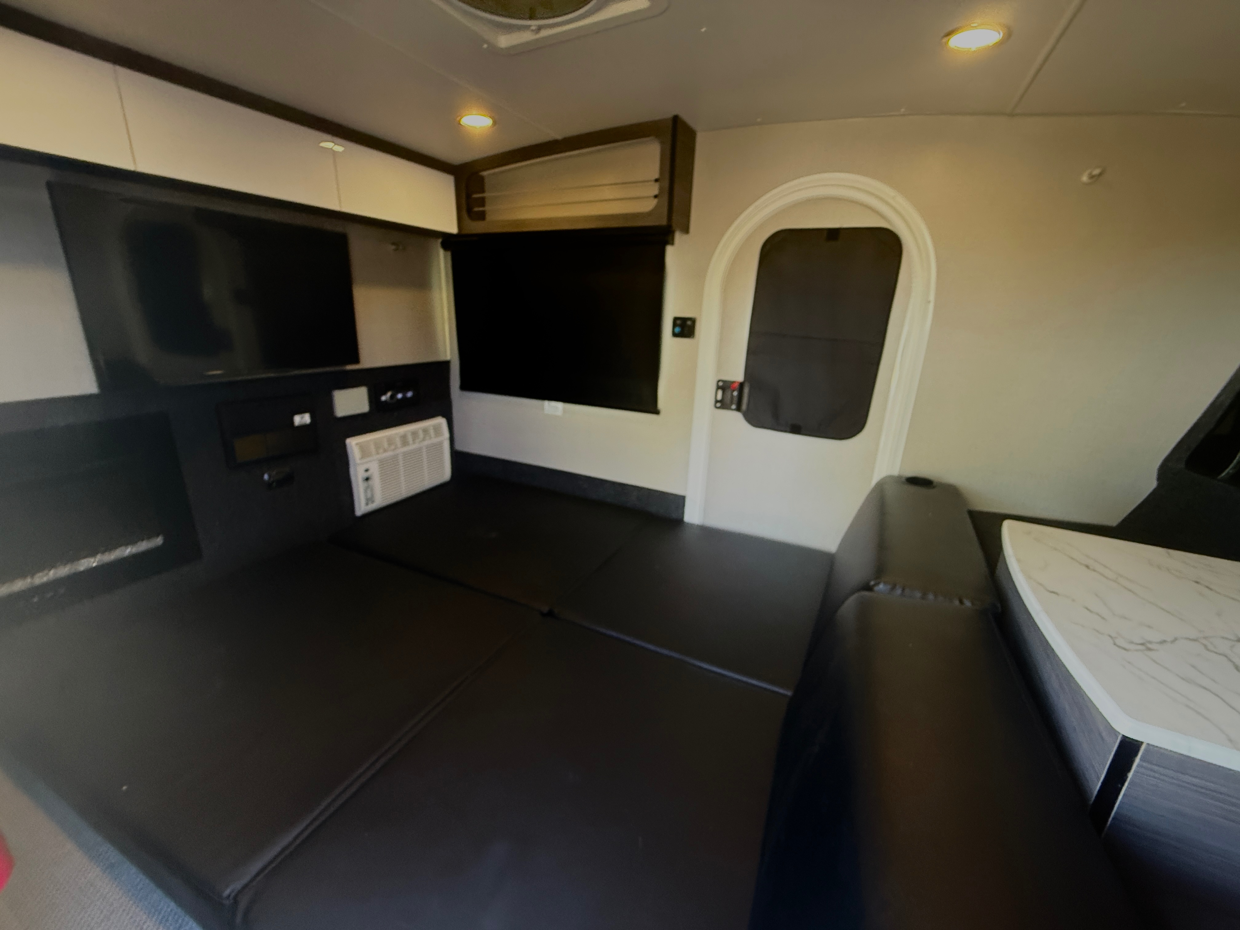 Cozy RV rental interior with a convertible bed, dual TVs, AC unit, and a small table. Bright overhead lights illuminate the space.. InTech RV Luna 2022