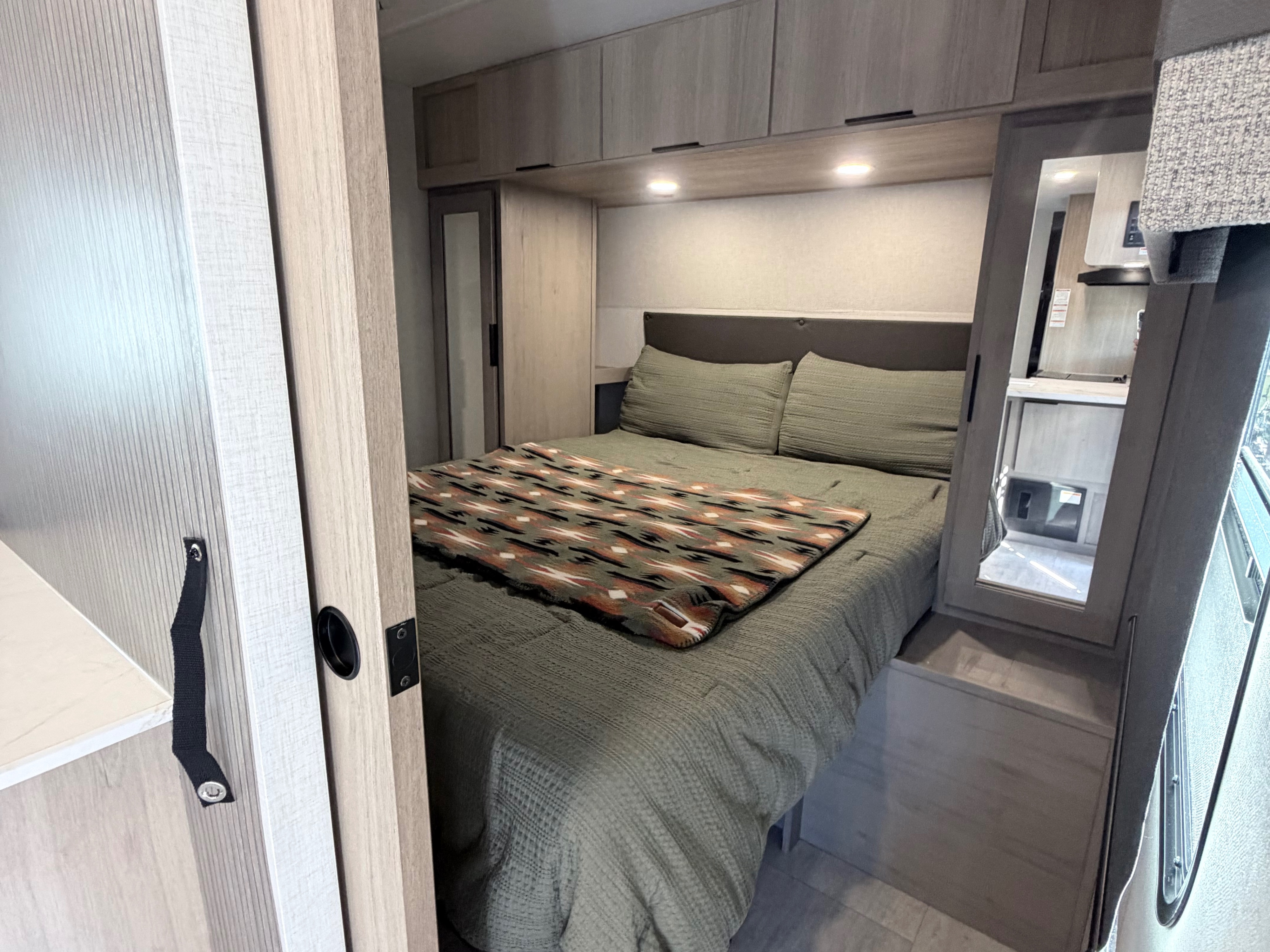 Cozy RV rental bedroom featuring a sage green bed, overhead cabinets with lighting, and a southwestern patterned blanket.. Dutchmen Kodiak 2026