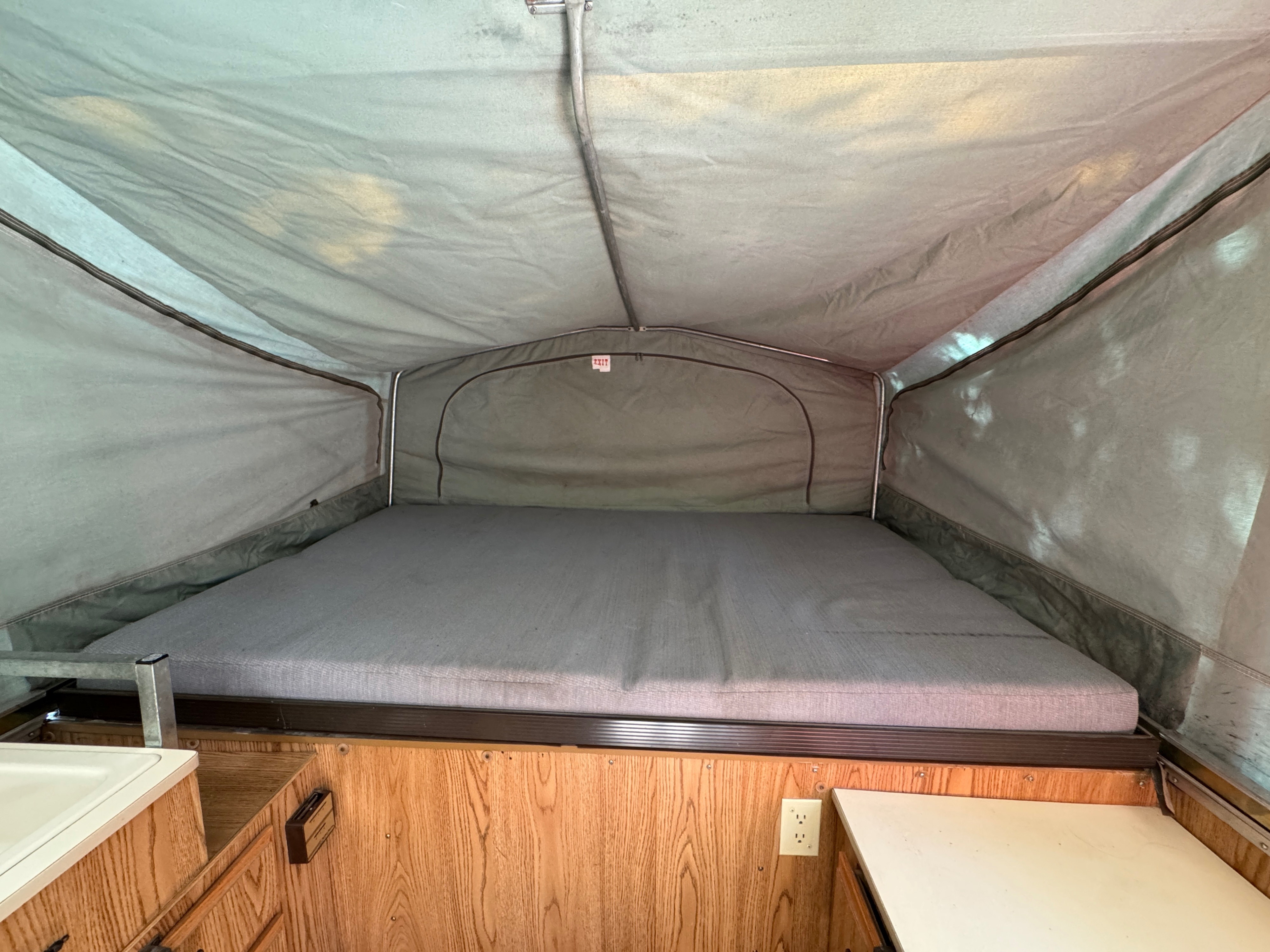 Spacious RV rental pop-up camper interior with a gray mattress, fabric walls, wood cabinetry, and an outlet. Ready for camping.. Jayco Jay Thrush 6 1989