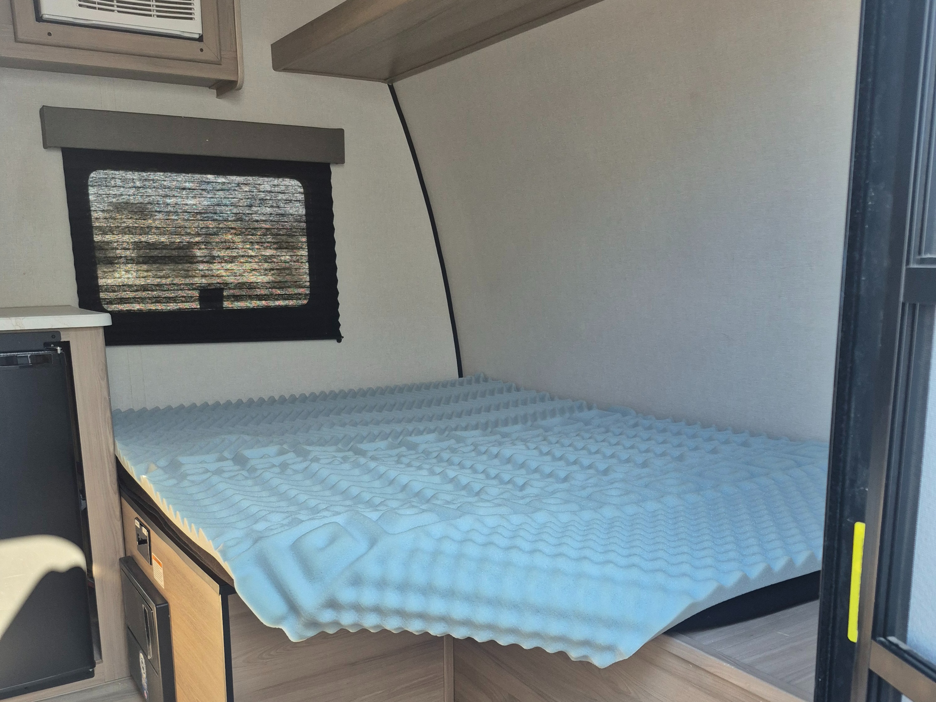 RV rental interior: a compact sleeping area with a blue egg crate mattress topper, window with blind, and cabinet.. Keystone RV 13B 2025