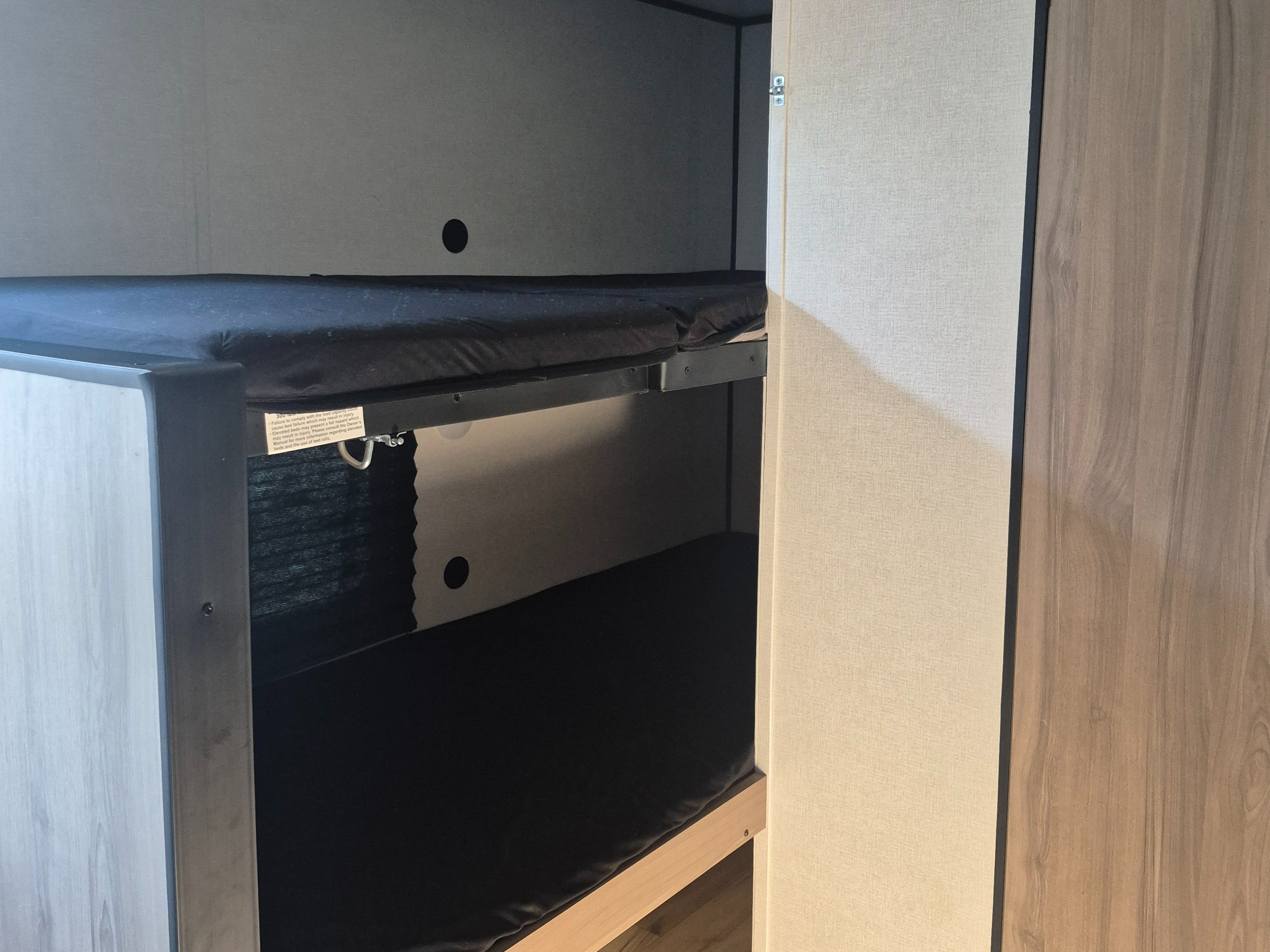RV rental interior showing bunk beds. Dark mattresses on metal frames with a safety warning label on the upper bunk.. Keystone RV 13B 2025