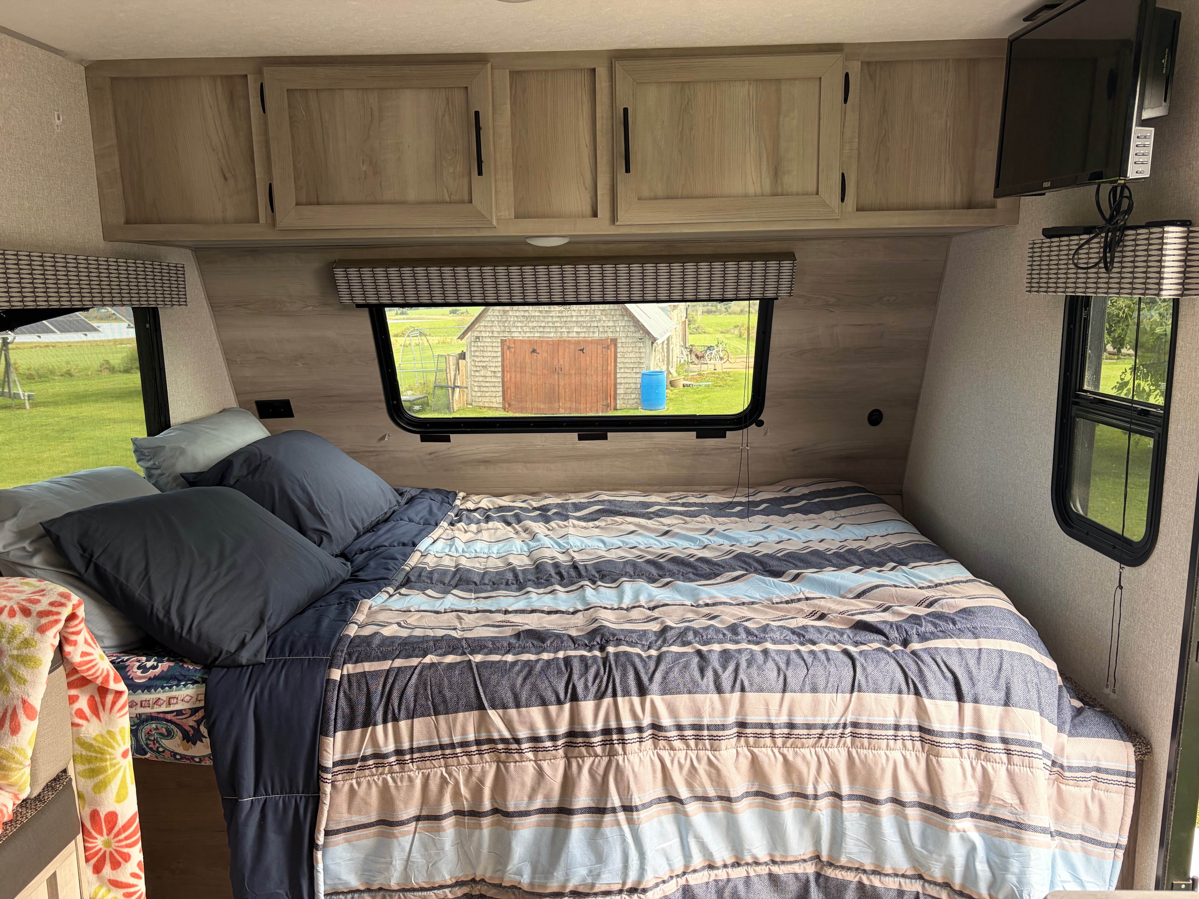 Cozy RV rental bedroom featuring a striped bed, overhead cabinets, TV, and a scenic window view of a shed and yard.. Sportsmen Classic 2022
